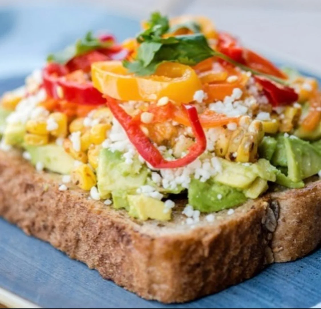 Open-faced sandwich with avocado, corn, diced vegetables, crumbled cheese, and sliced peppers on toasted bread