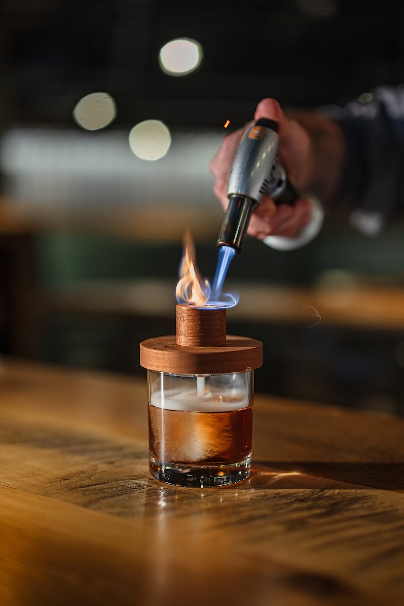 A person performing a flame test on a wooden lid over a glass container filled with a dark liquid, using a handheld blowtorch at night.
