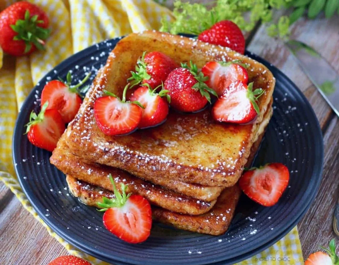 Plate of French toast topped with strawberries and powdered sugar, served on a dark plate with a yellow checkered cloth underneath.