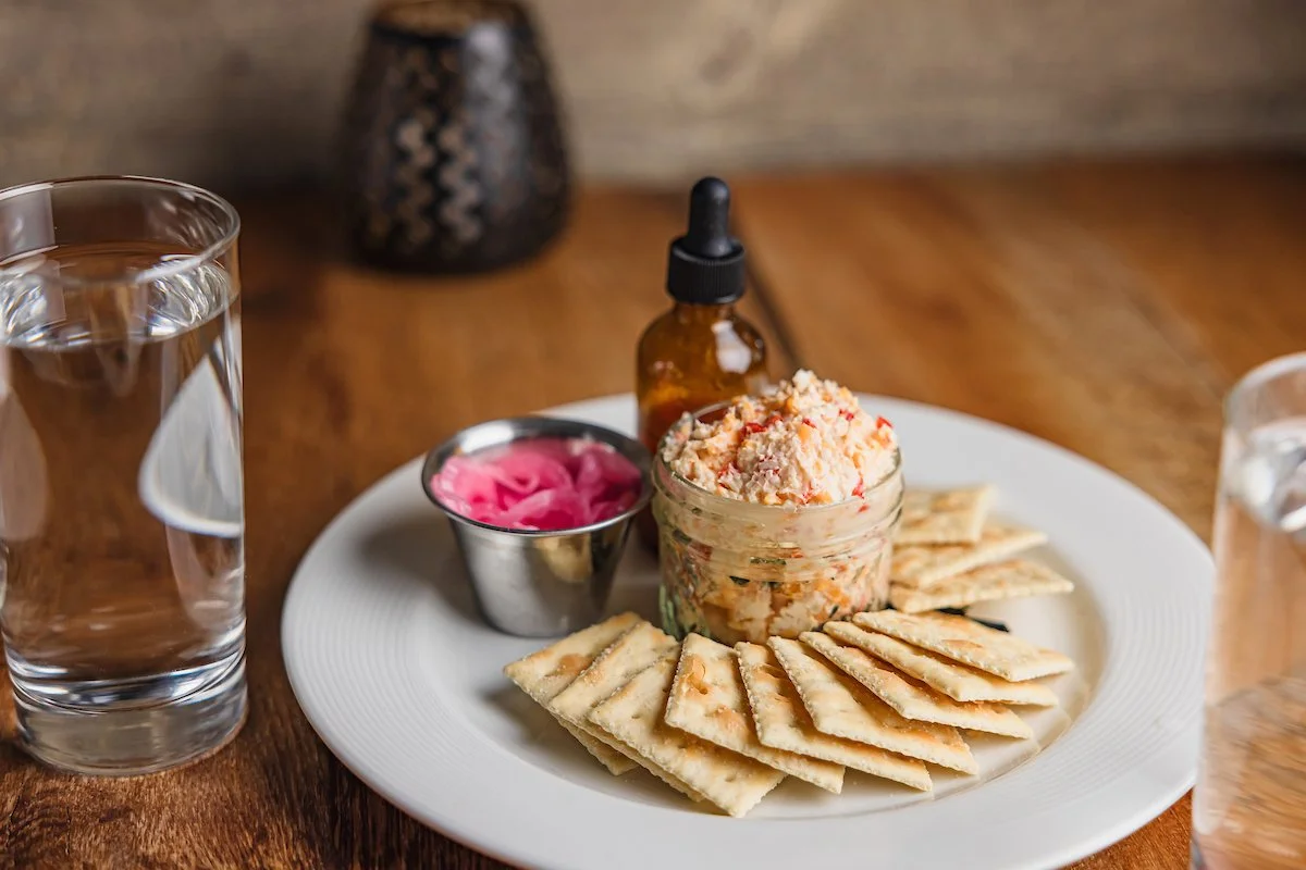 Plate of crackers with a jar of seafood spread, pickled onions, hot sauce, and two glasses of water.
