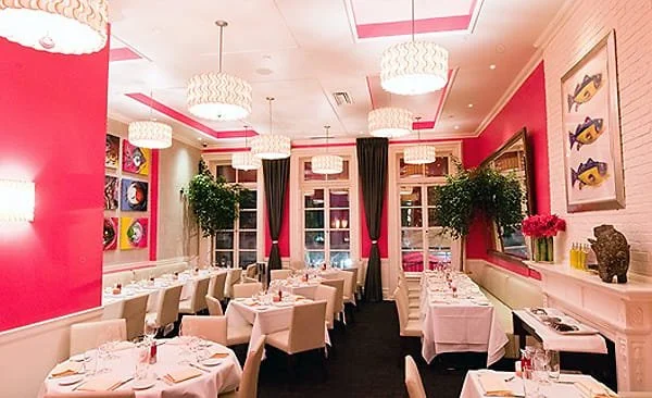 Elegant restaurant with pink walls, white tablecloths, and modern chandeliers, decorated with framed fish artwork and potted plants.