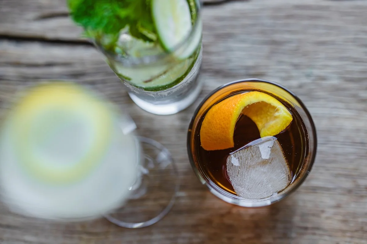 Three drinks on a wooden surface, one with a lemon wedge and ice, one with cucumber slices and ice, and one with a lemon wedge and ice.