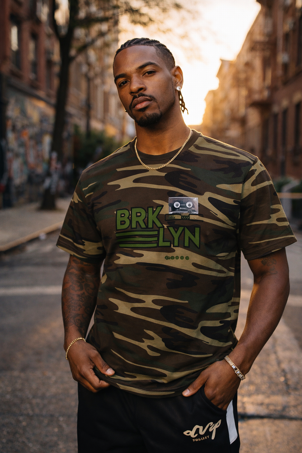 A young man with braided hair and tattoos, wearing a camouflage T-shirt with green text and a cassette tape design, standing on a city street during sunset.