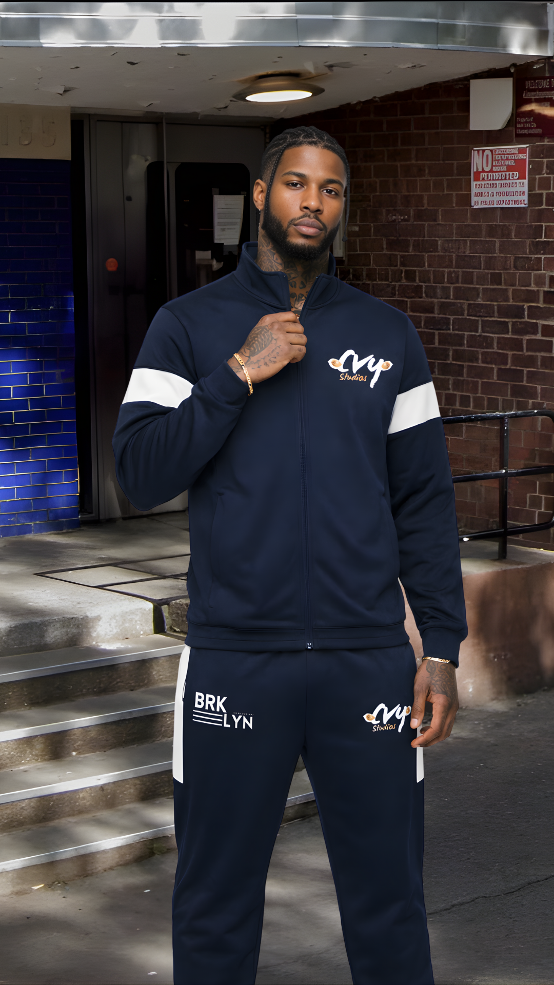 A man standing outdoors at night wearing a navy blue tracksuit with white accents and logos, with a urban background.