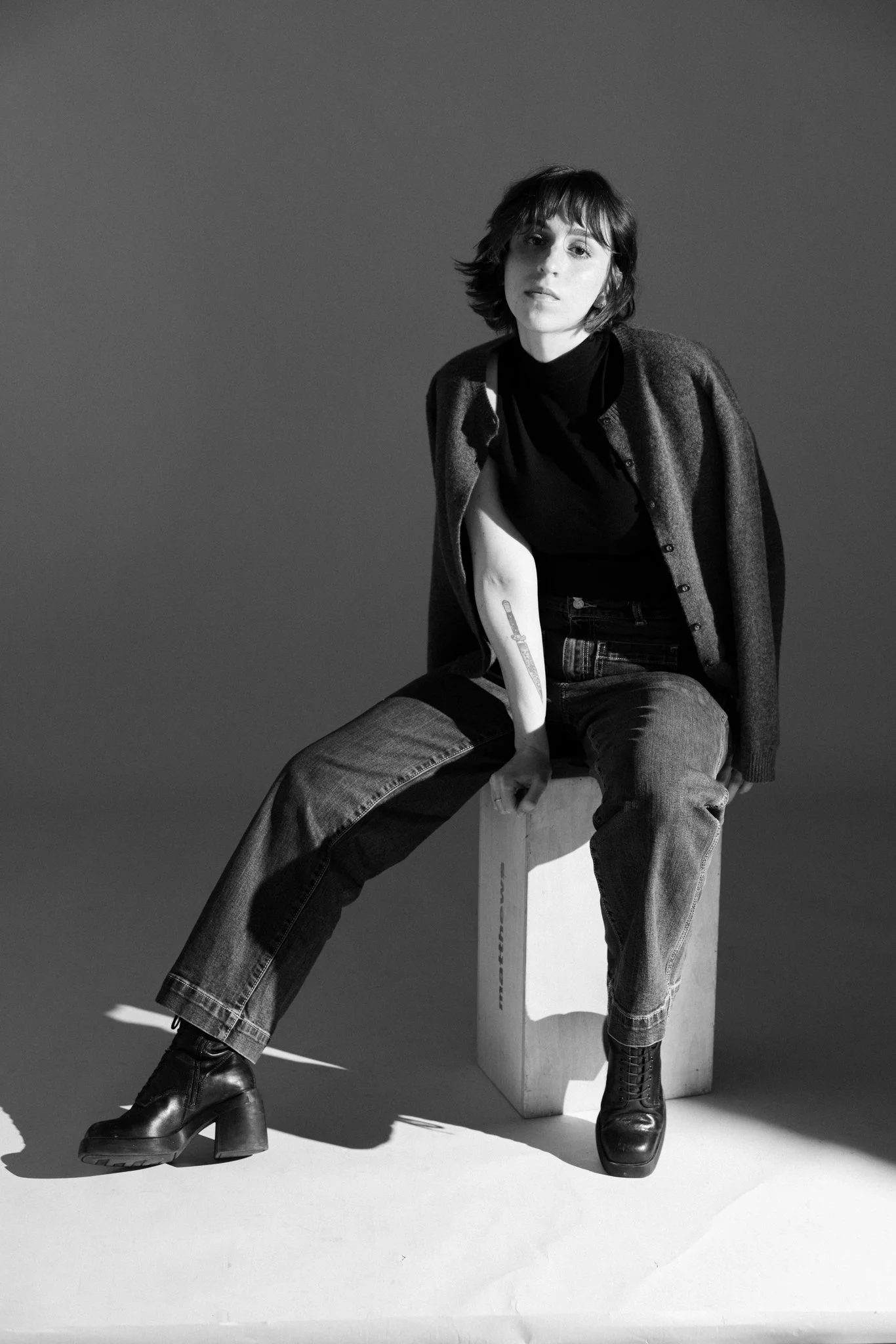 Black and white photo of Cleo Shine with chin-length hair sitting on a stool against a plain background. She is wearing a dark turtleneck, a checkered coat draped over her shoulders, and high-heeled boots, with a tattoo on her left arm.