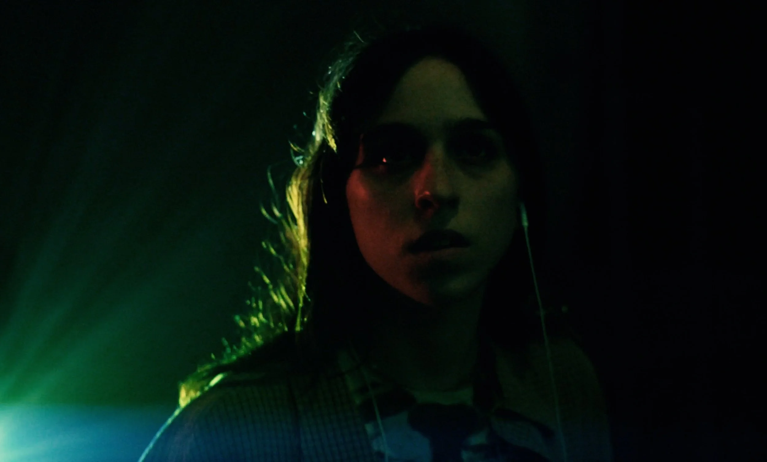 A woman with dark hair wearing sunglasses and earphones, illuminated by green and red lighting in a dark setting.