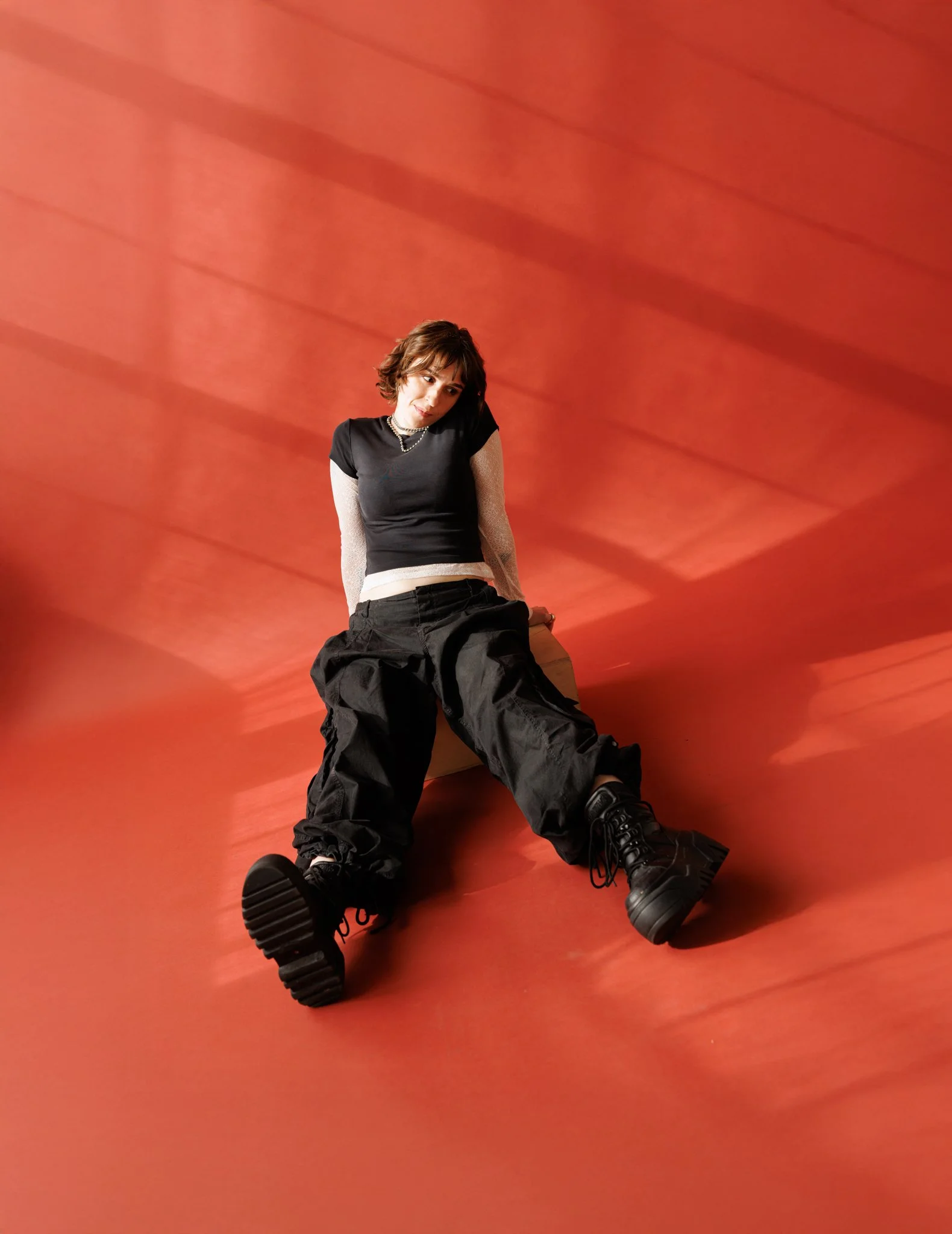 Cleo Shine with short brown hair, wearing a black T-shirt layered over a white long-sleeve shirt, black pants, and black boots, sitting on the floor against an orange background with diagonal shadow lines.