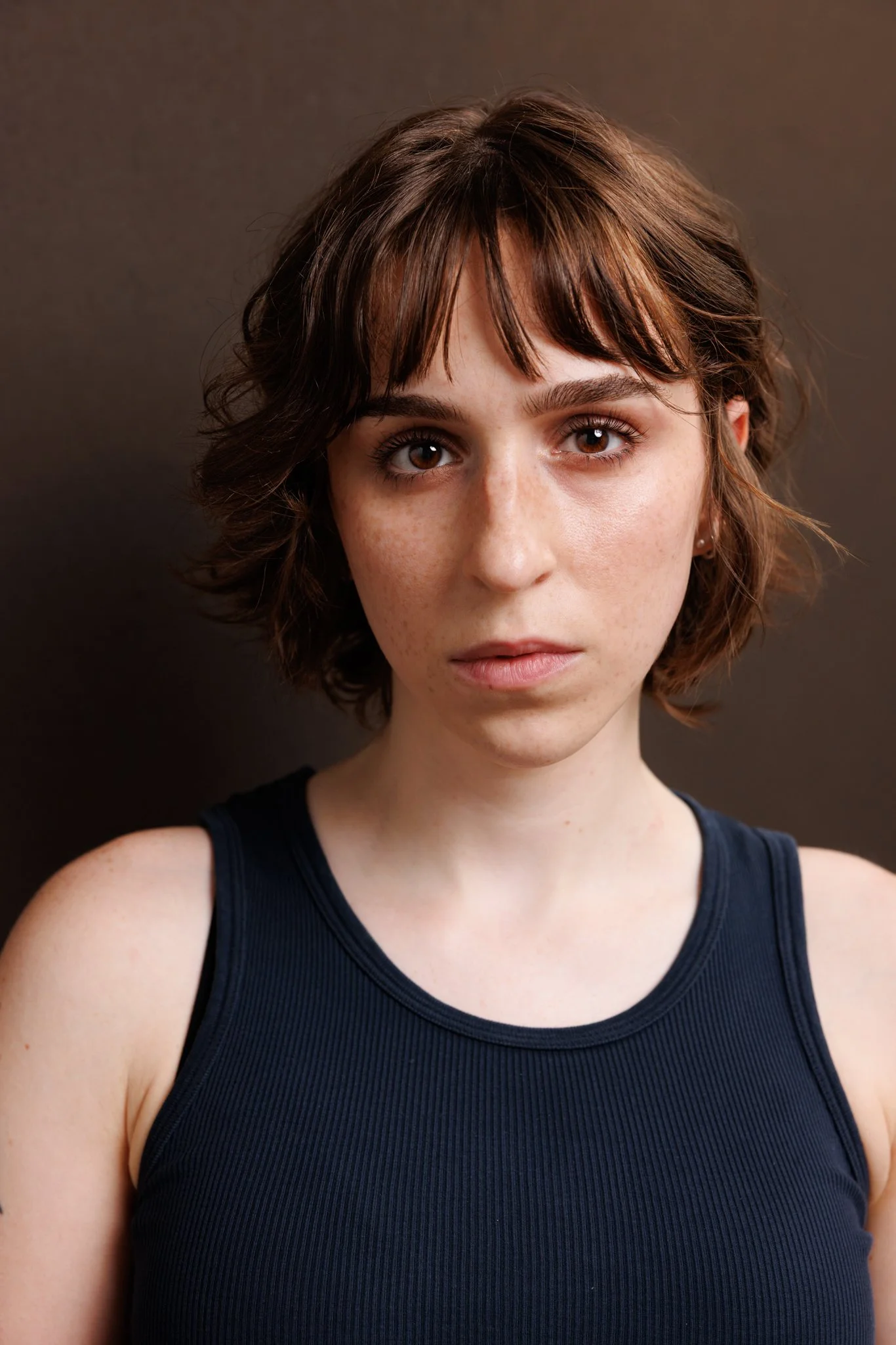 Actress Cleo Shine with short, wavy brown hair wearing a black sleeveless top, against a dark background.
