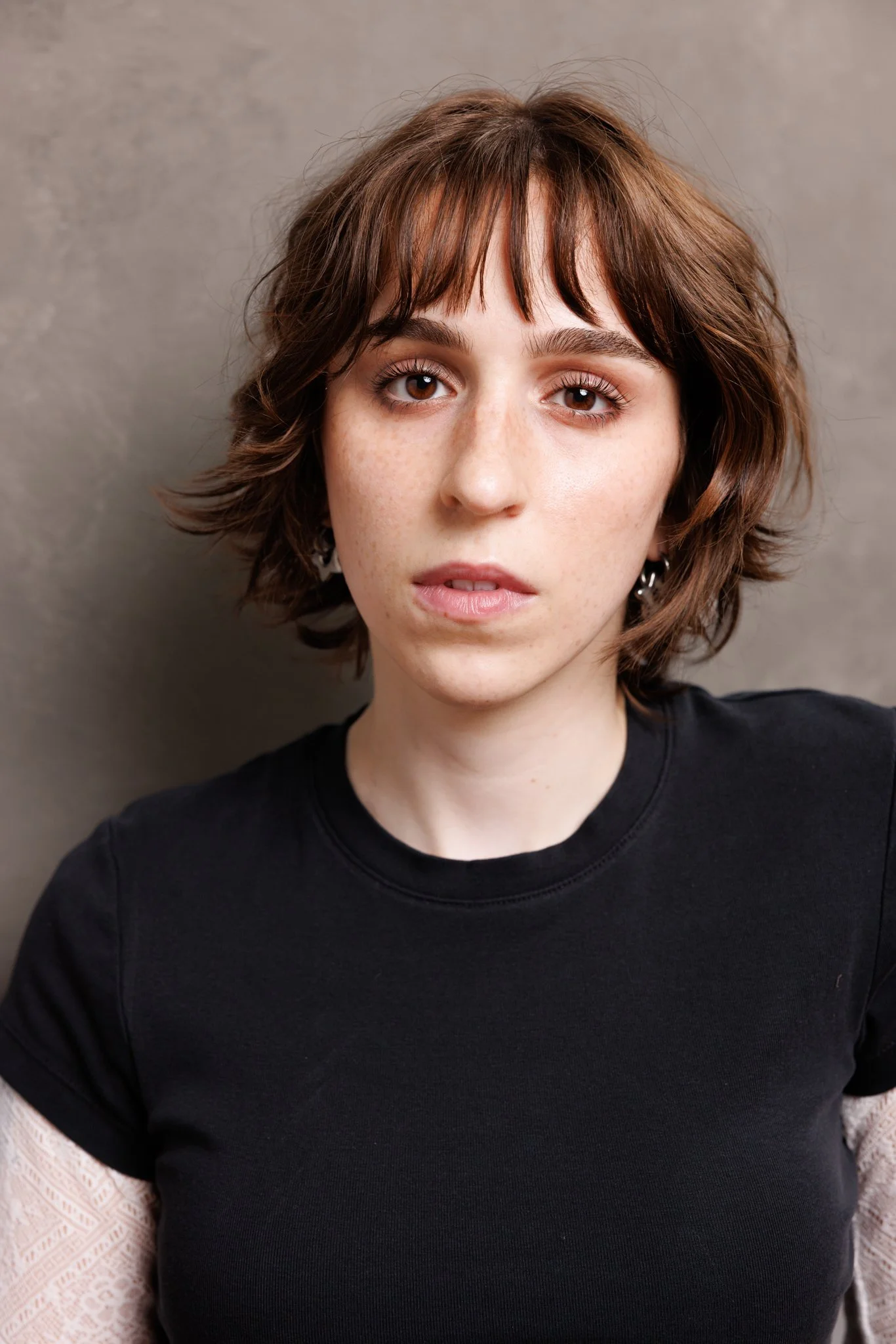 Actress Cleo Shine with light brown hair and brown eyes, wearing a black top and earrings, against a gray background.