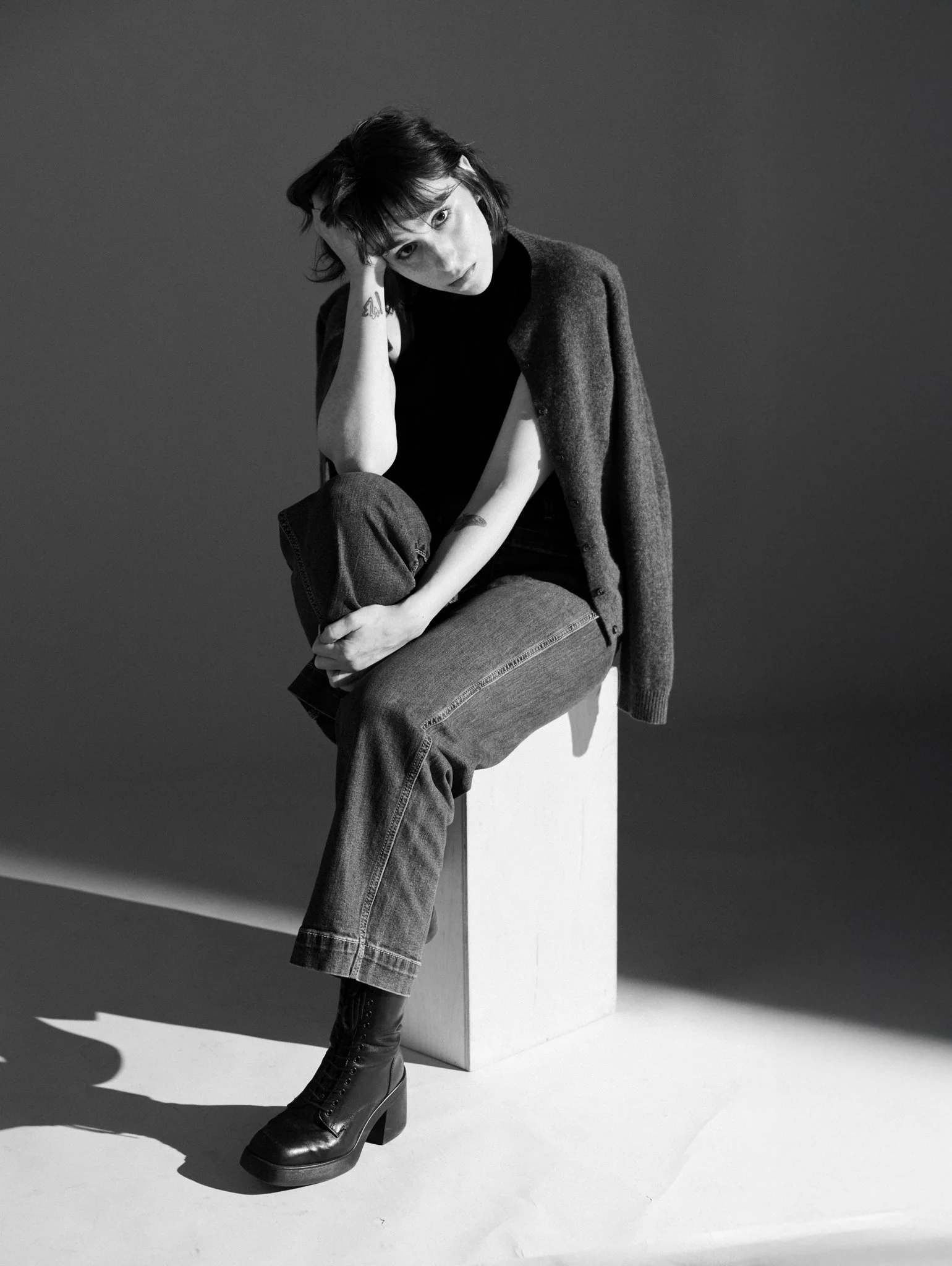A black and white photo of Cleo Shine with dark hair, sitting on a white cube, wearing a dark top, long coat, ripped jeans, and black platform boots, leaning her head on her hand, with a plain dark background.