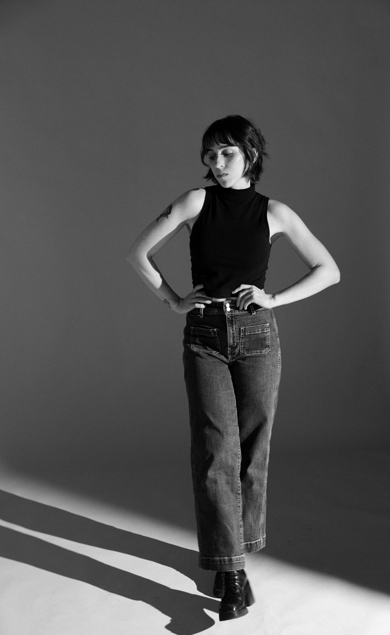 Cleo Shine with short, wavy hair, wearing a sleeveless black top, high-waisted jeans, and ankle boots, standing with one hand on her hip, against a plain background with a shadow cast on the floor.