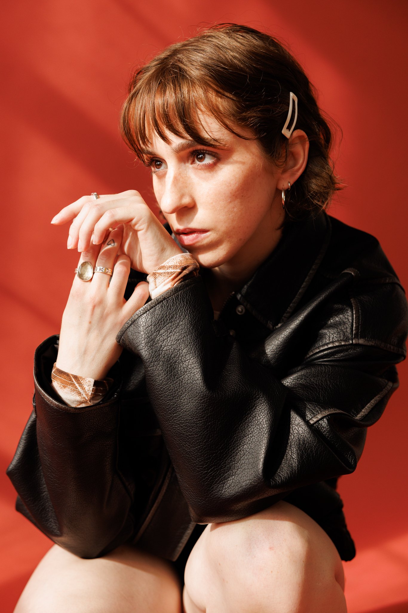 Cleo Shine with short brown hair and freckles, wearing hoop earrings and a beige hair clip, sitting with her knee close to her face, wearing a black leather jacket and a patterned shirt underneath, against a red background.