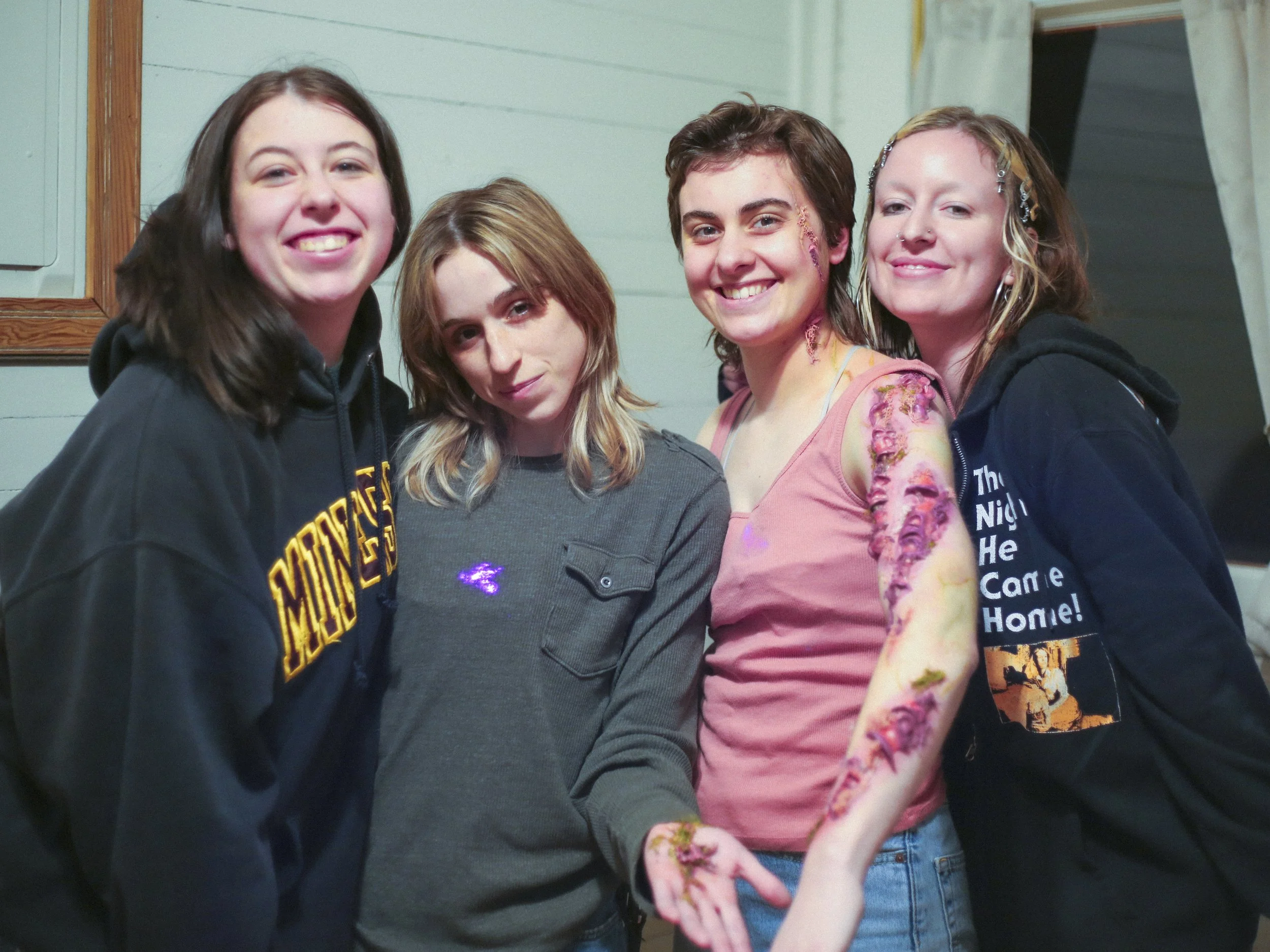 A group of five young women standing closely together and smiling for a photo indoors, with wooden wall paneling and a mirror in the background.