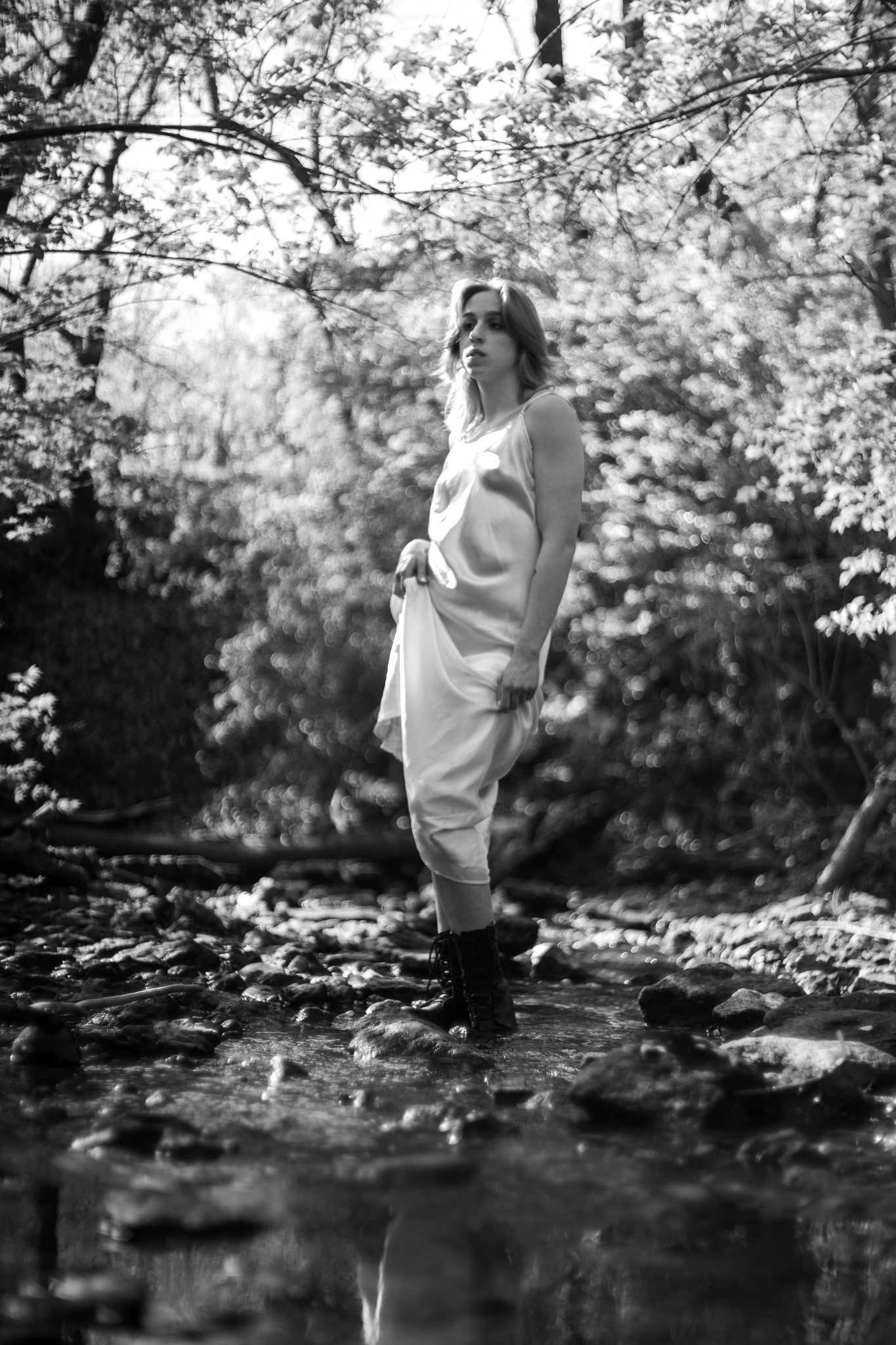 Cleo Shine standing in a stream in a forest, wearing light-colored clothing and dark boots.