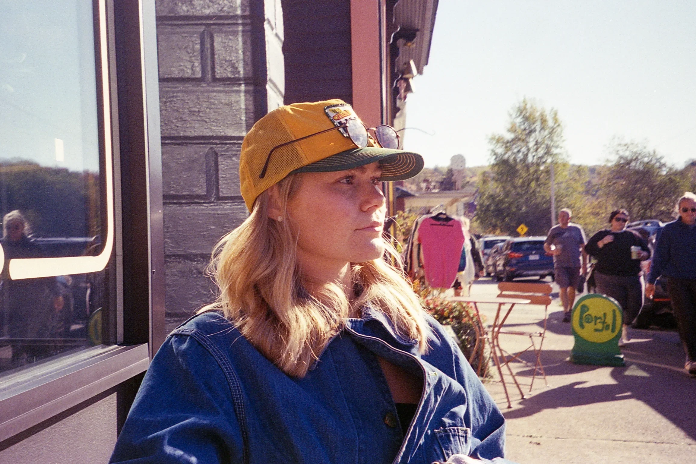 A woman with long blonde hair, wearing a yellow cap with sunglasses on top, a dark denim jacket, sitting outdoors in a cafe or market area with pedestrians and parked cars in the background.