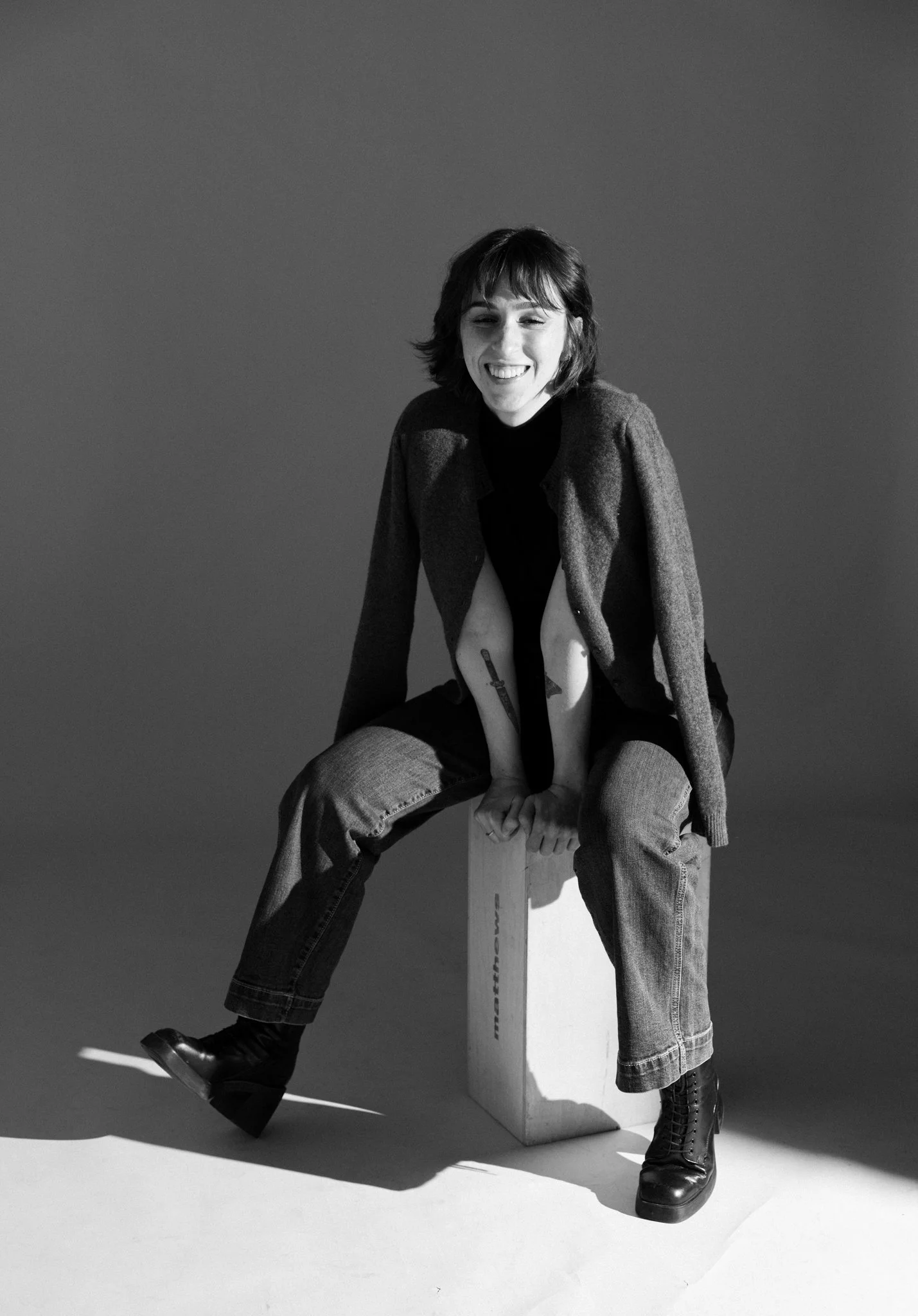 Black and white photo of Cleo Shine smiling, sitting on a box, wearing a blazer, jeans, and boots, with an uneven haircut and tattoos on her arms, against a plain background.