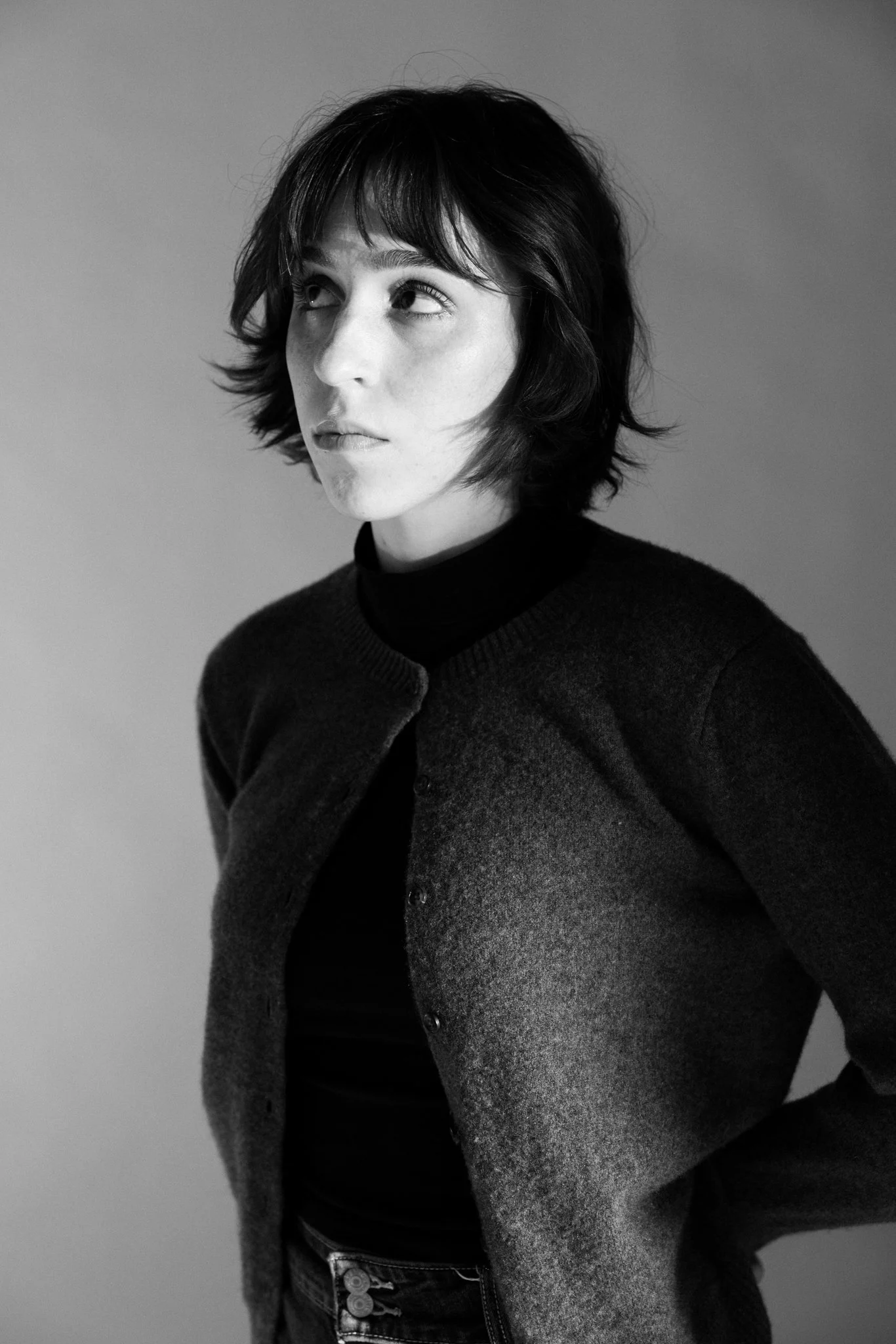 Black and white portrait of Cleo Shine with short, dark, layered hair, wearing a dark turtleneck and a buttoned cardigan, looking upward to her right.
