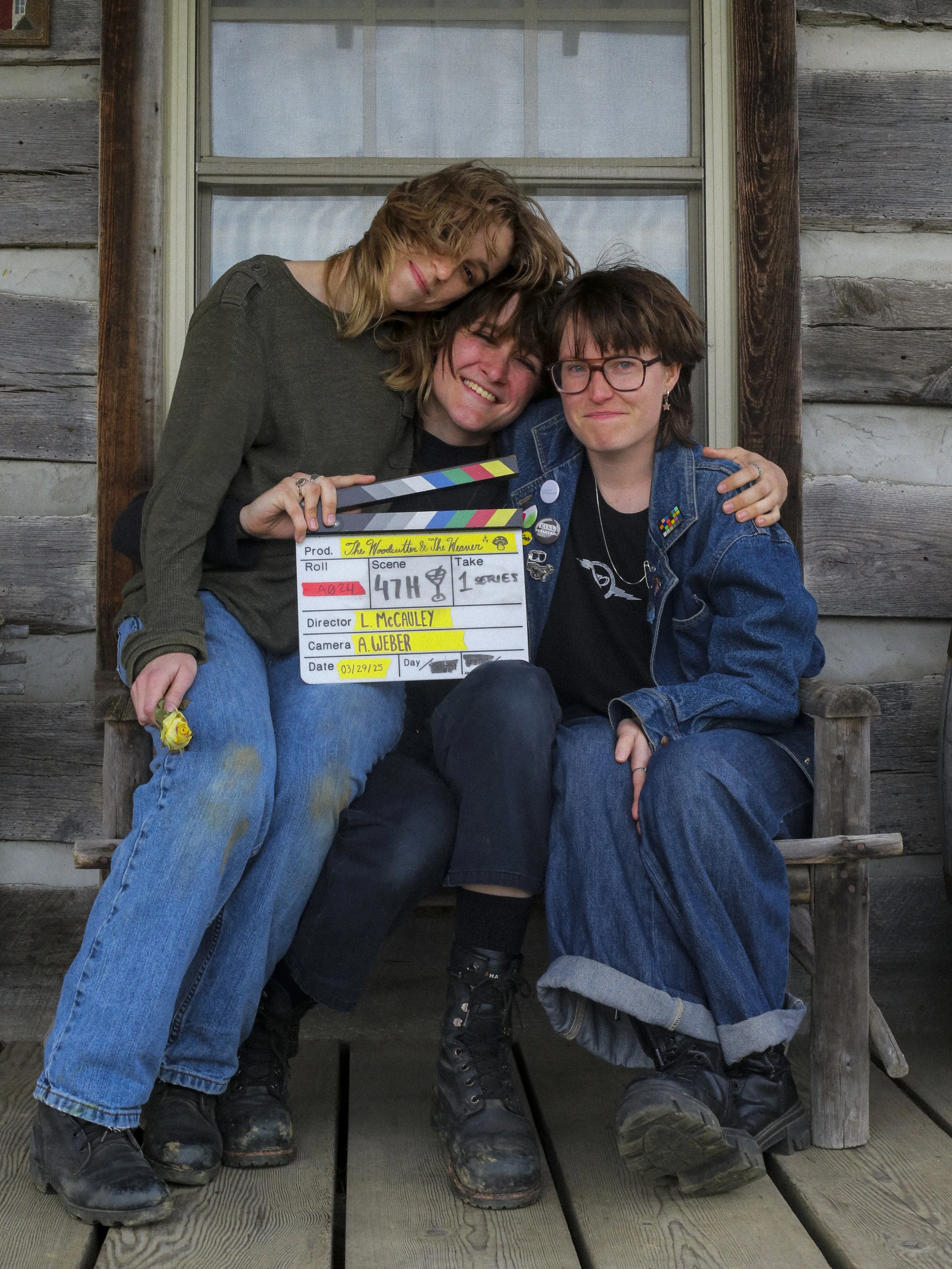 Lily McCauley, Cleo Shine, and Audrey Weber wrapping The Woodcutter & The Weaver, sitting on a wooden porch, holding a film slate, smiling, with a rustic house in the background.