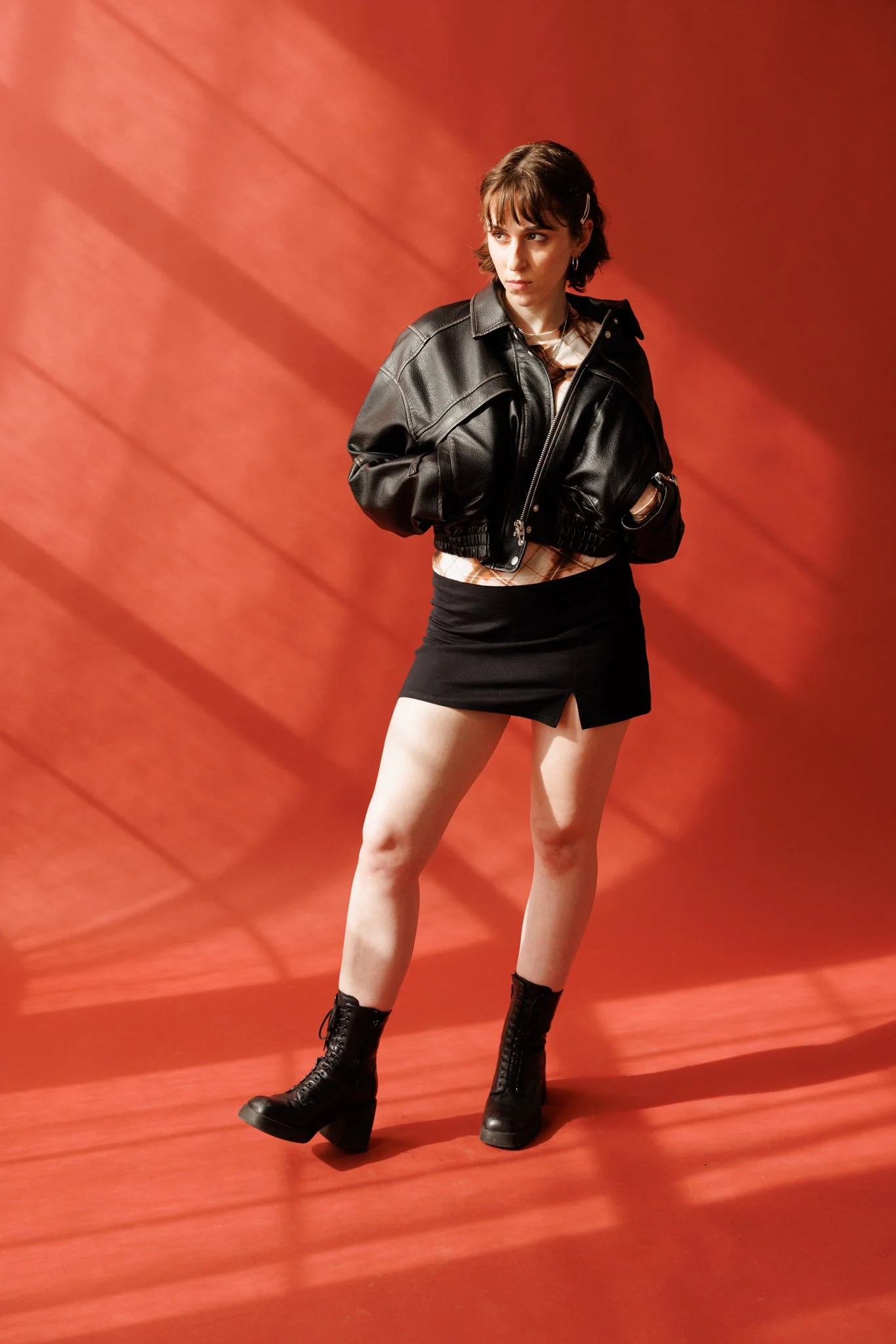 Cleo Shine stands in front of an orange background with dramatic shadows. She is wearing a black leather jacket, a beige plaid skirt, and black platform boots. Her expression is confident and contemplative.