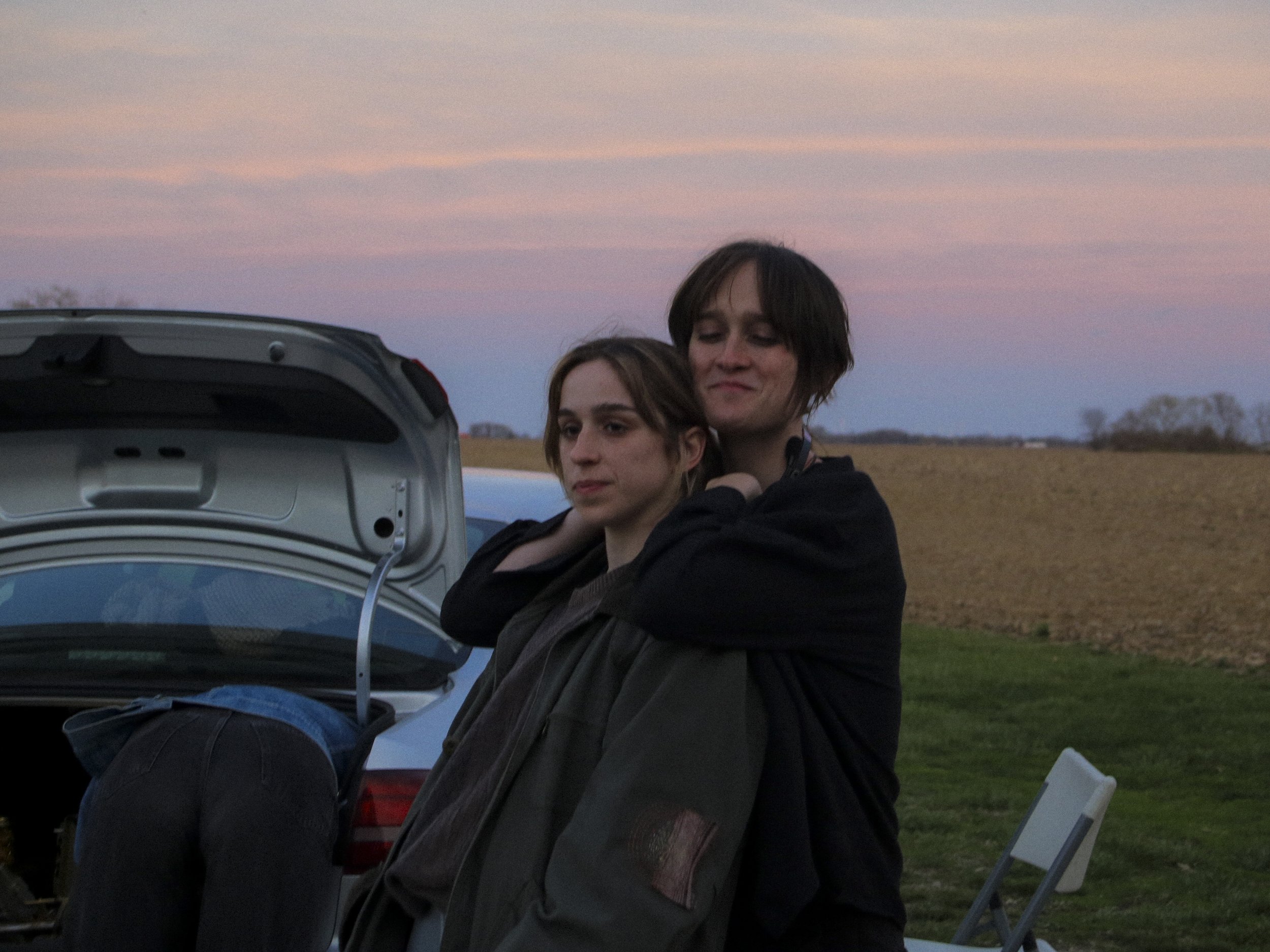 Two people with dark hair, one with shorter hair and the other with longer hair, stand close together near the open trunk of a car during sunset or sunrise in a rural area, with fields and trees in the background.