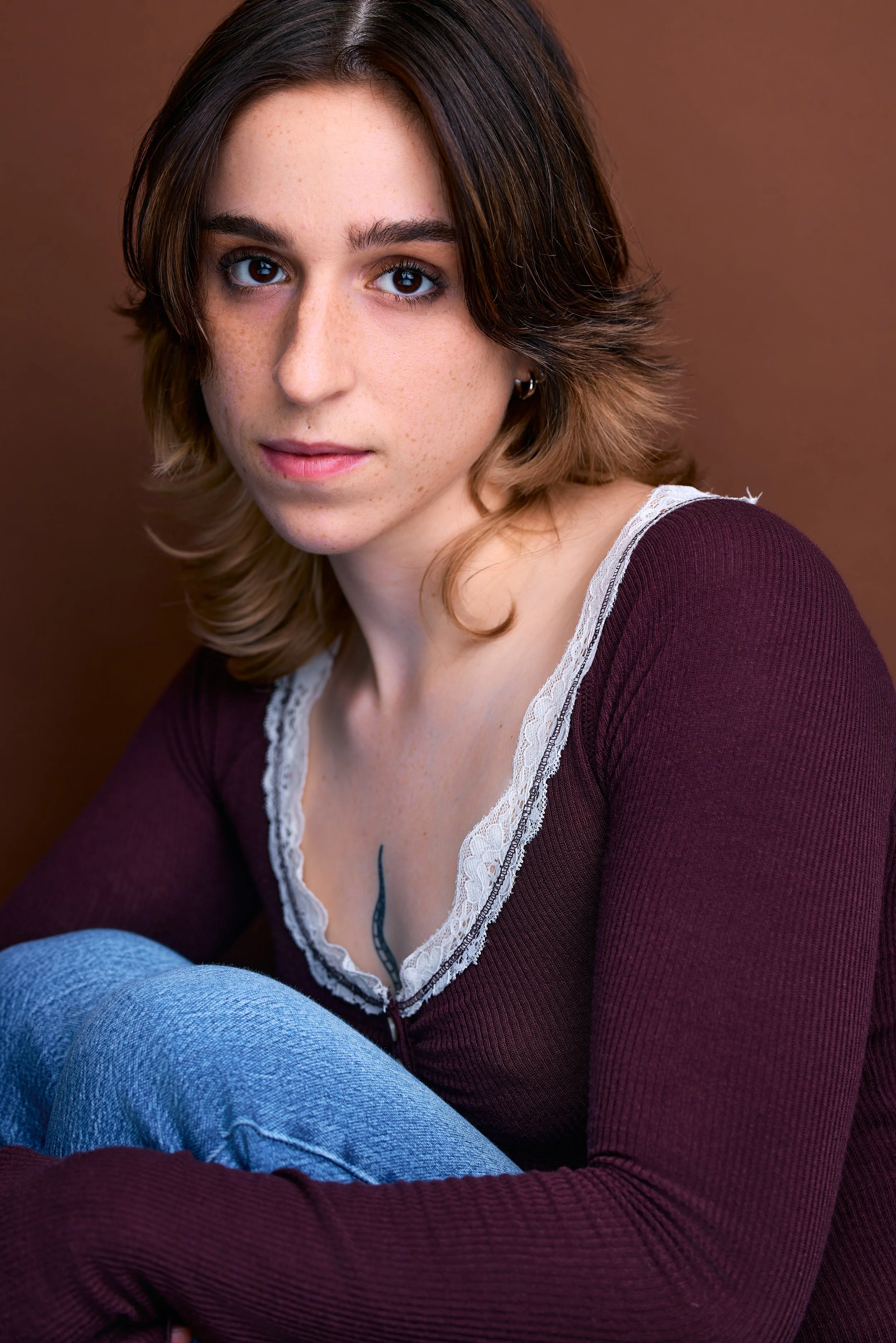 Close-up portrait of Cleo Shine with short brown hair, blue eyes, and freckles, wearing a maroon top with lace details and light blue jeans, against a brown background.