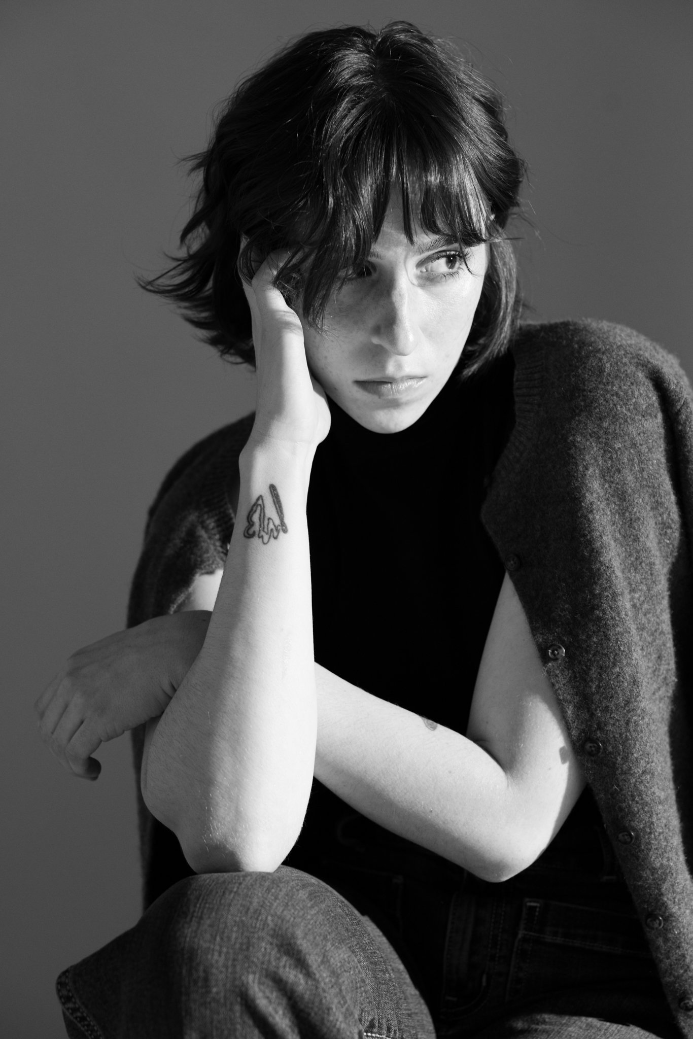 Cleo Shine with short dark hair and light skin, wearing a dark turtleneck and a cardigan, resting her head on her hand with tattoos on her forearm, sitting with her arms crossed in a black-and-white photo.