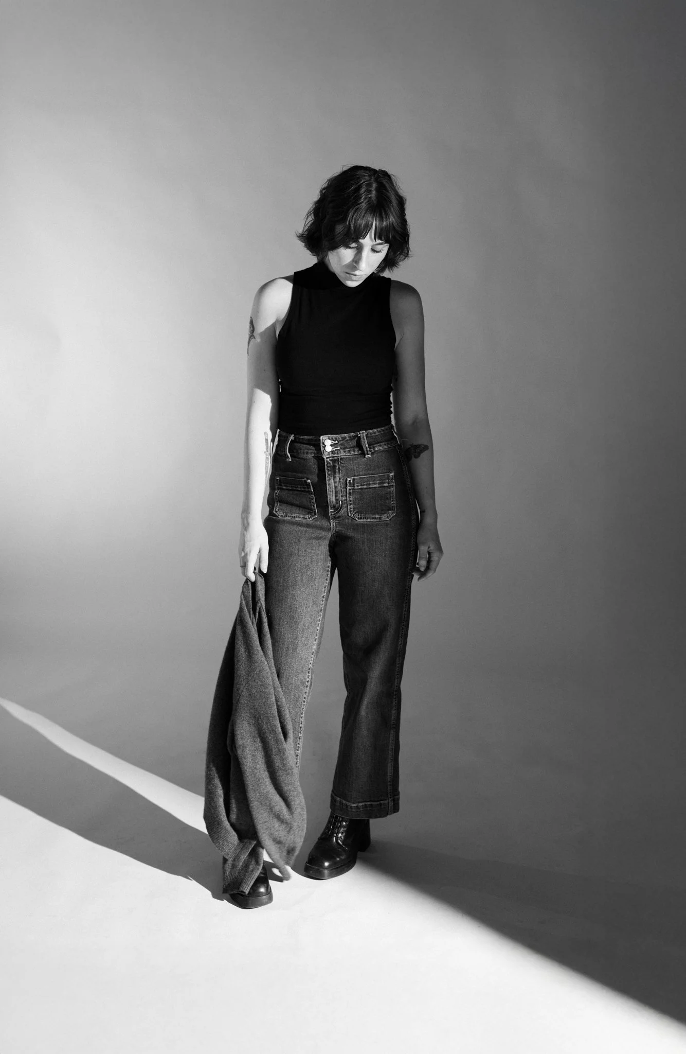 Black and white photo of Cleo Shine with dark wavy hair wearing a sleeveless black turtleneck, wide-leg jeans, and lace-up boots, standing against a plain gray background. She is looking down with her head slightly bowed.