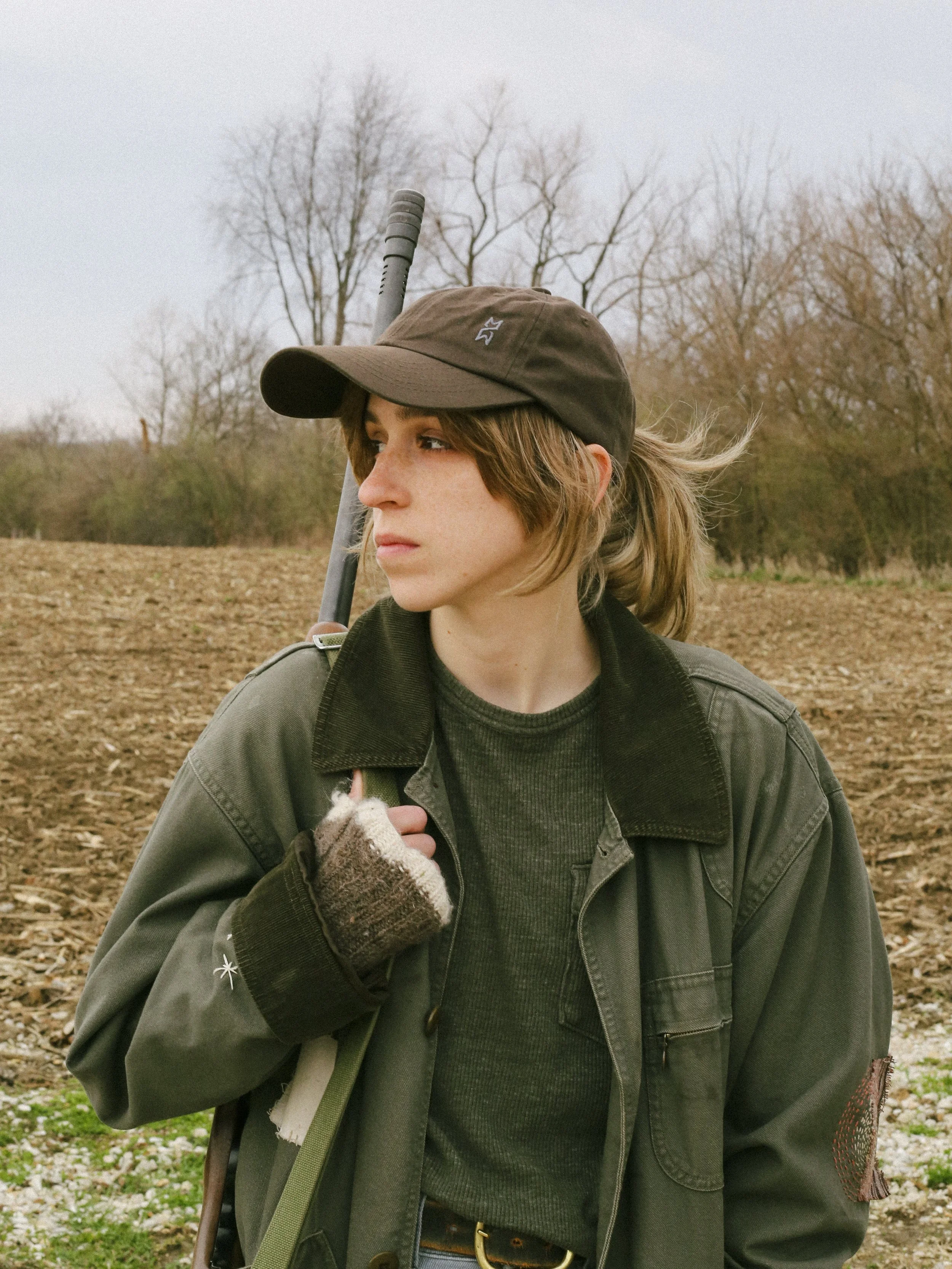 Cleo Shine with light brown hair and fair skin, wearing a brown cap, a dark green jacket, and fingerless gloves, stands outdoors in a field with leafless trees in the background.