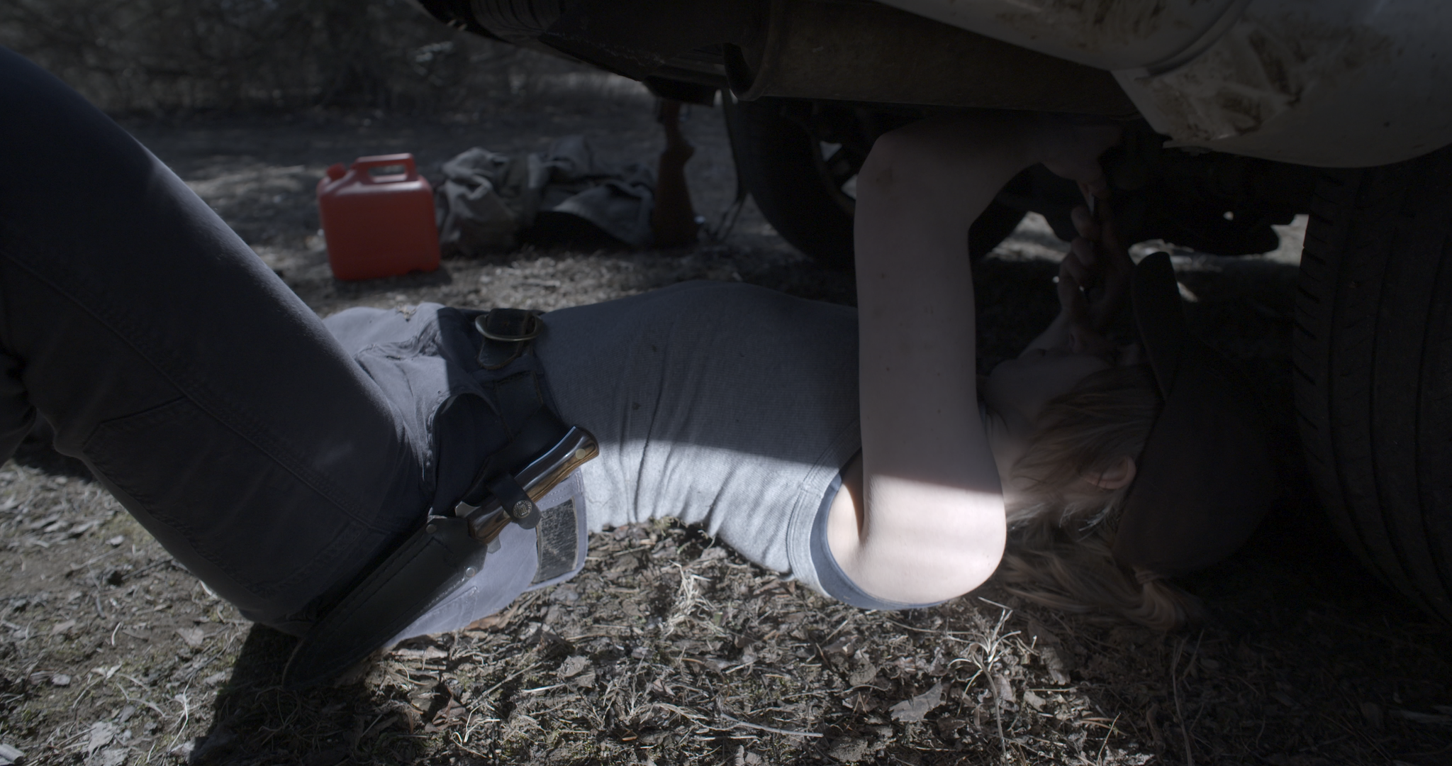 A person lying underneath a vehicle, working on its underside, with tools and supplies nearby on the dirt ground.