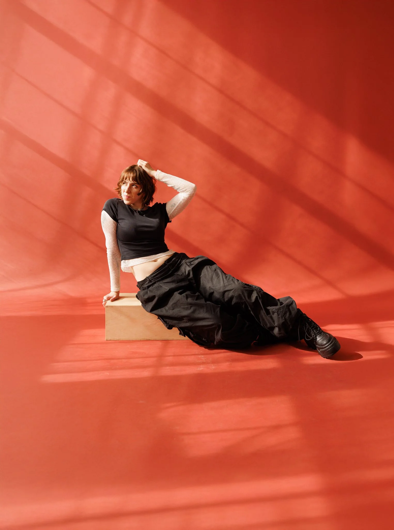 Cleo Shine with brown hair and casual clothing sitting on a wooden box against a red background with diagonal shadows.