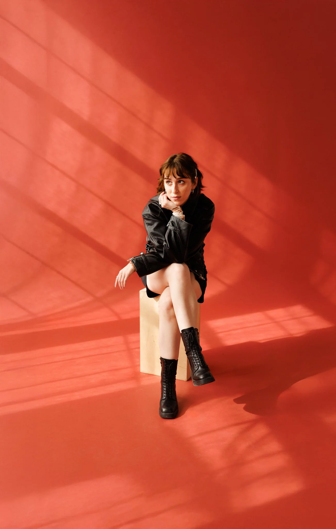 Cleo Shine sitting on a beige block against a red wall with shadows and diagonal lines, wearing a black leather jacket, black shorts, and lace-up black combat boots. She is resting her chin on her hand and looking to the side.