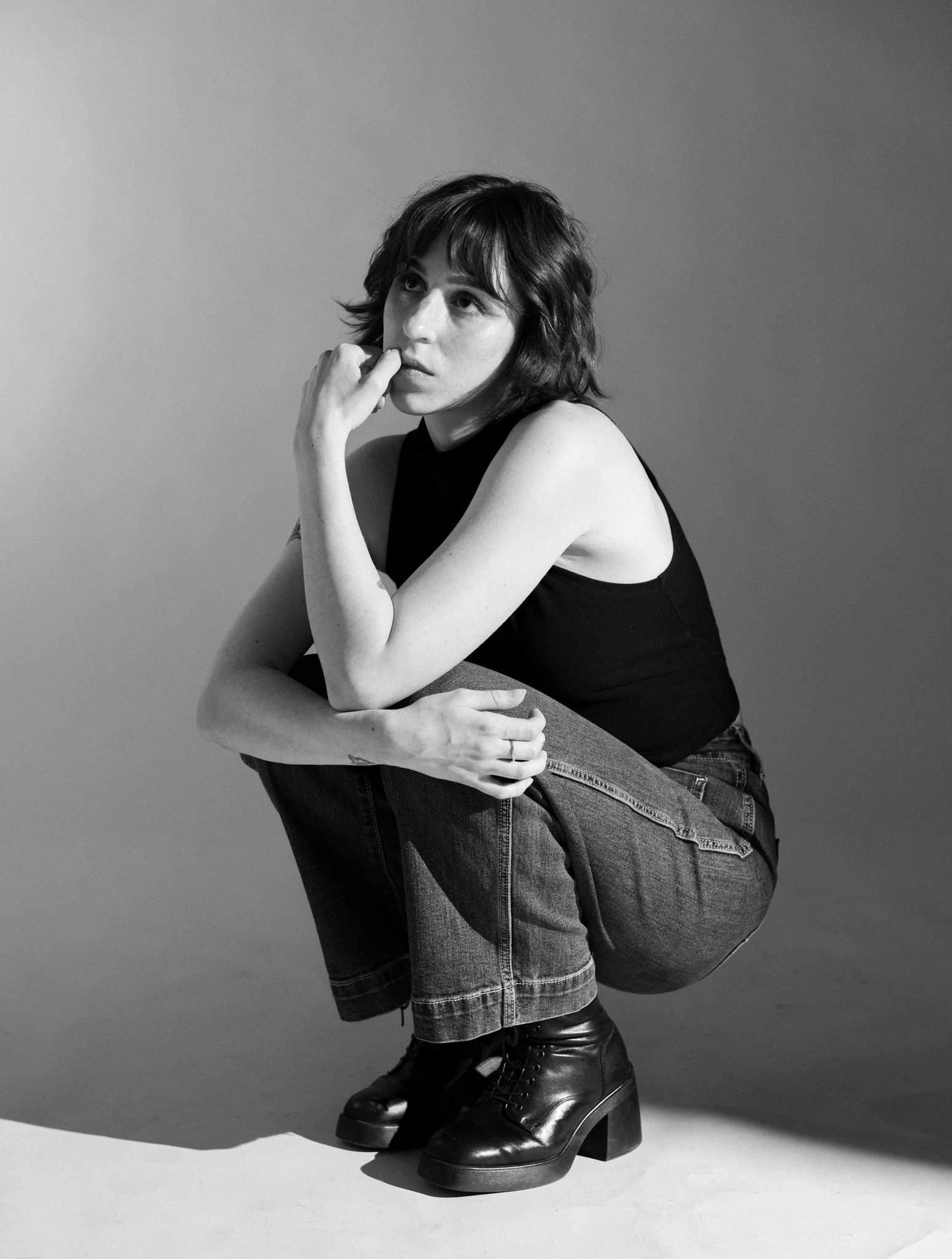 Cleo Shine with short wavy hair sitting on a chair with one knee raised. She is wearing a sleeveless top, jeans, and chunky leather boots. She rests her chin on her hand, looking thoughtfully to the side against a plain background.