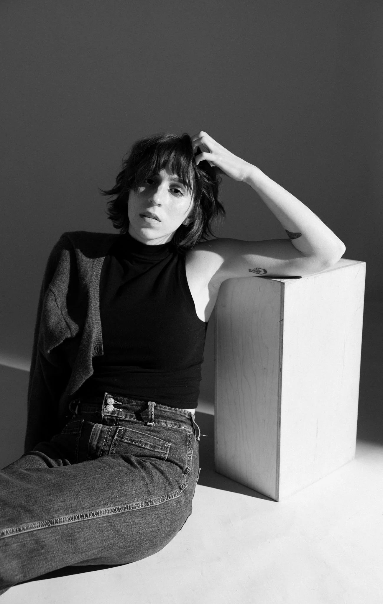 Cleo Shine wearing a sleeveless top and high-waisted jeans, sitting on the floor with her back against a wall and leaning on a tall rectangular block. She has one arm raised, with her hand on her head, and appears to be looking at the camera.