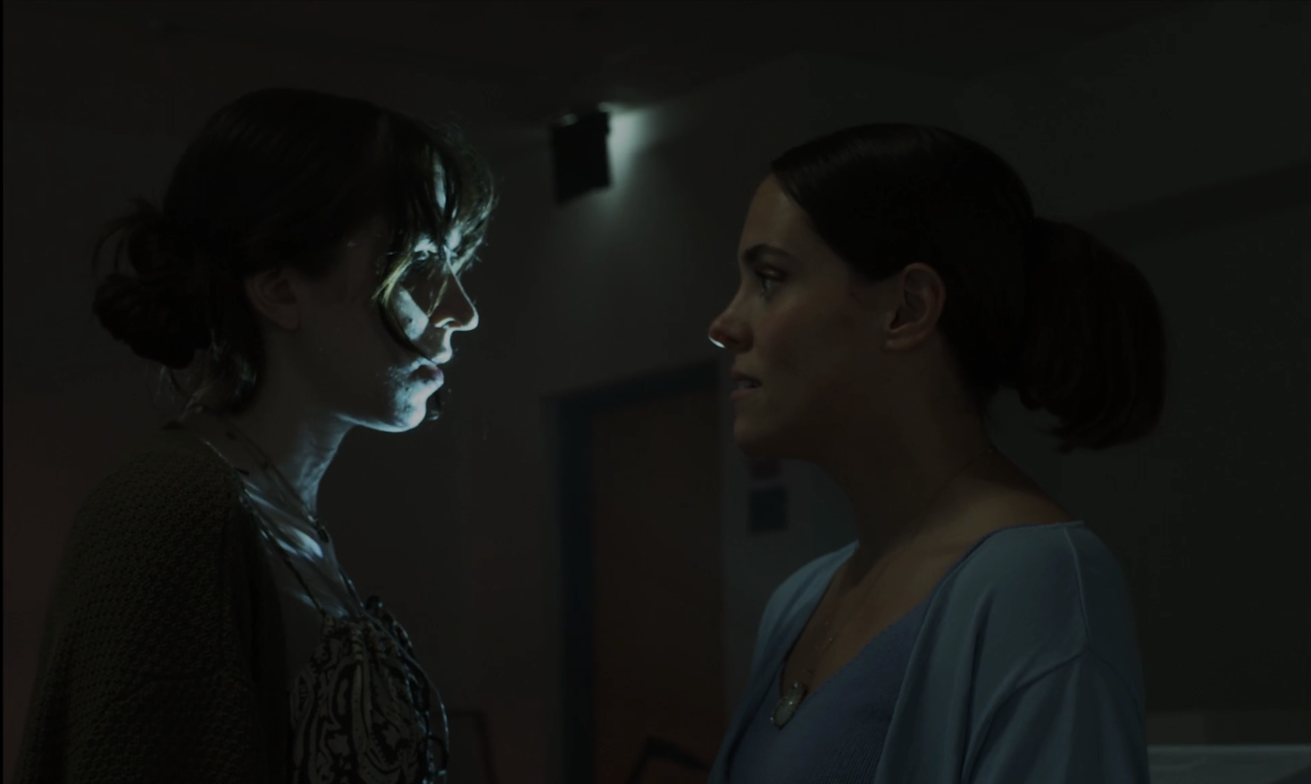 Two women face each other in a dimly lit room, staring intensely at each other. One woman has wavy hair and is illuminated by a soft glow, while the other has straight hair tied back.