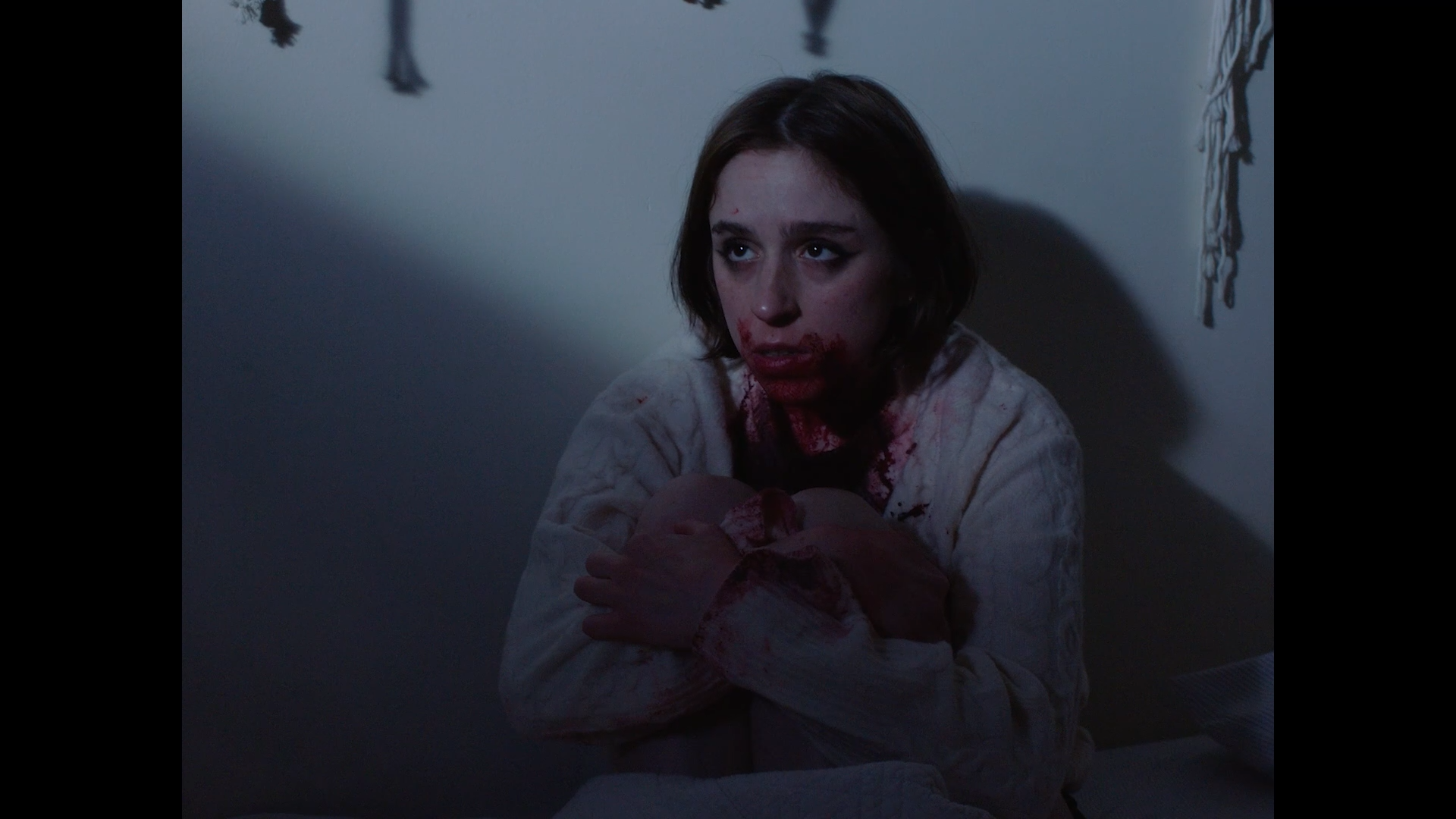 A woman with short brown hair, blood on her face, and wearing a bloodstained white sweater sitting on the floor with her arms crossed, looking upward in a dimly lit room.