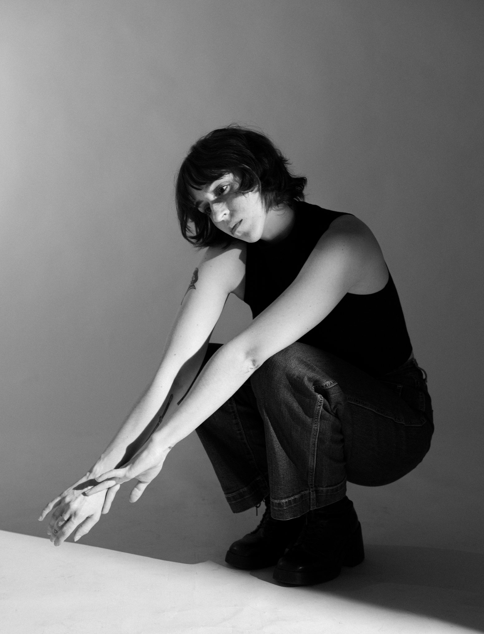 Cleo Shine with dark, shoulder-length hair, wearing a sleeveless black top and ripped jeans, is posed with her body bent forward and her arms extended downward, holding her hands together. 