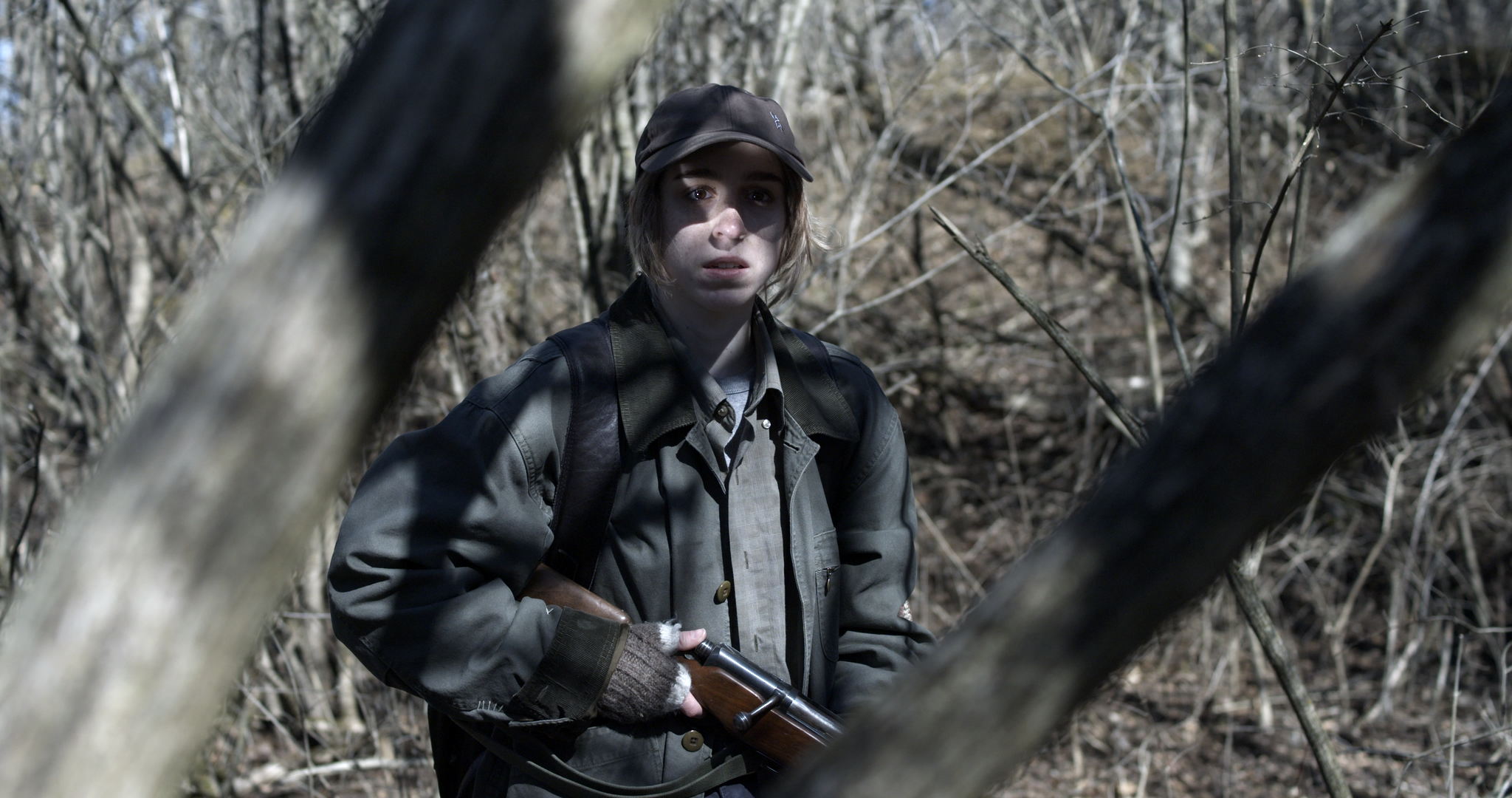 A young woman in camouflage clothes and a cap holding a rifle, standing in a forest with bare trees.