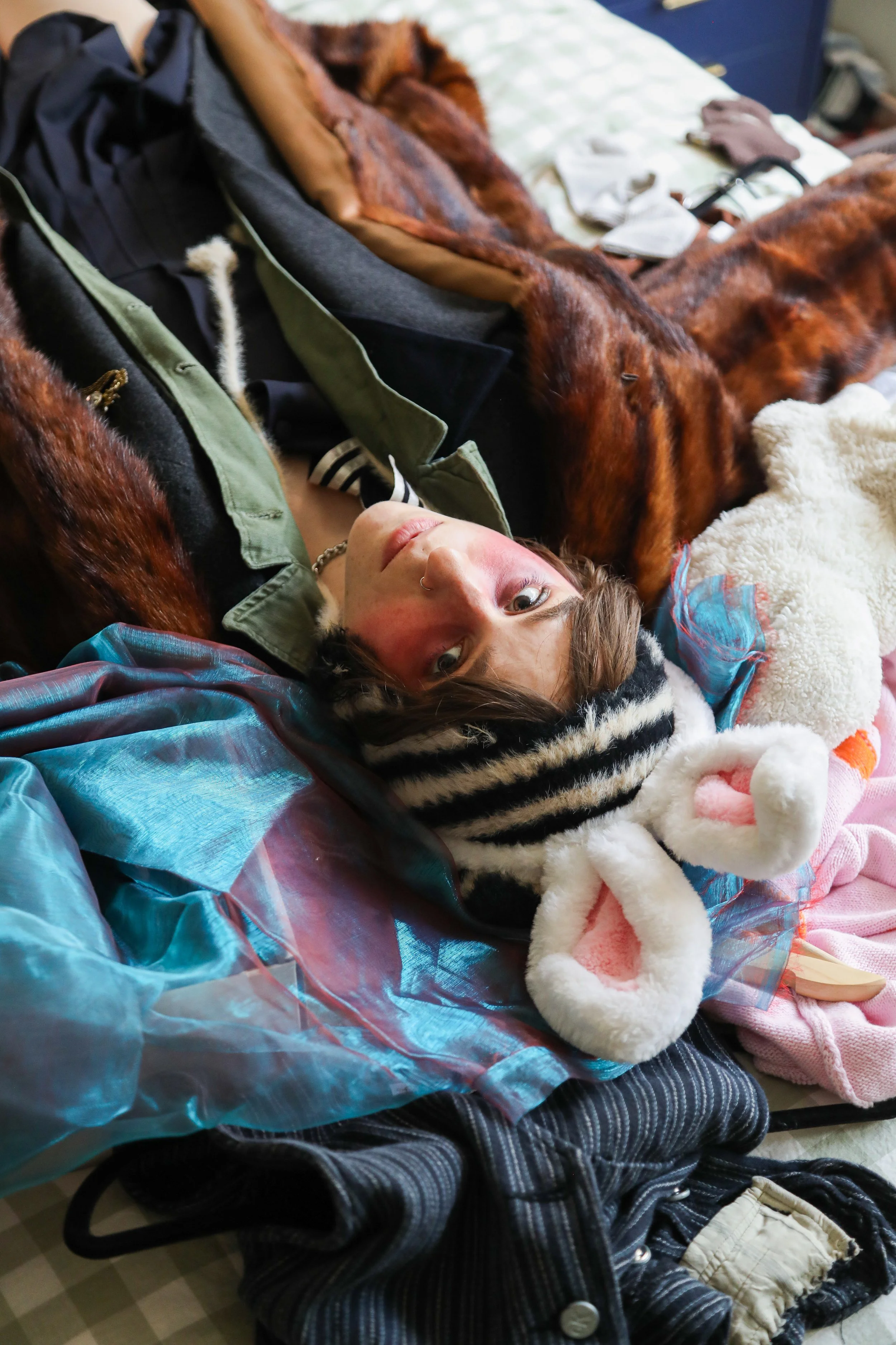 Cleo Shine as shot by Macgregor Salmon lying on a bed with assorted clothing and accessories around, wearing a striped jacket, beige shorts, and a faux fur coat, with a hood and details like rings and a necklace visible.