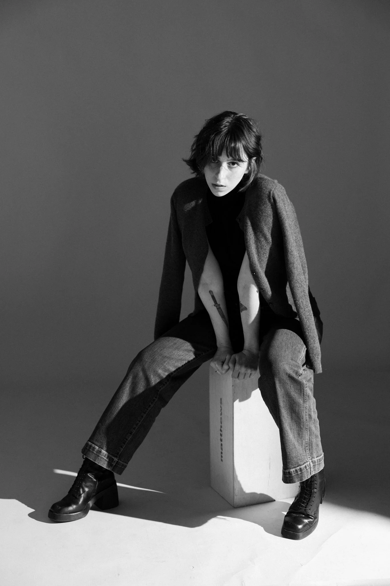 Black and white photo of Cleo Shine with short hair, sitting on a wooden block, wearing a coat, turtleneck, baggy jeans, and black chunky boots, looking at the camera.