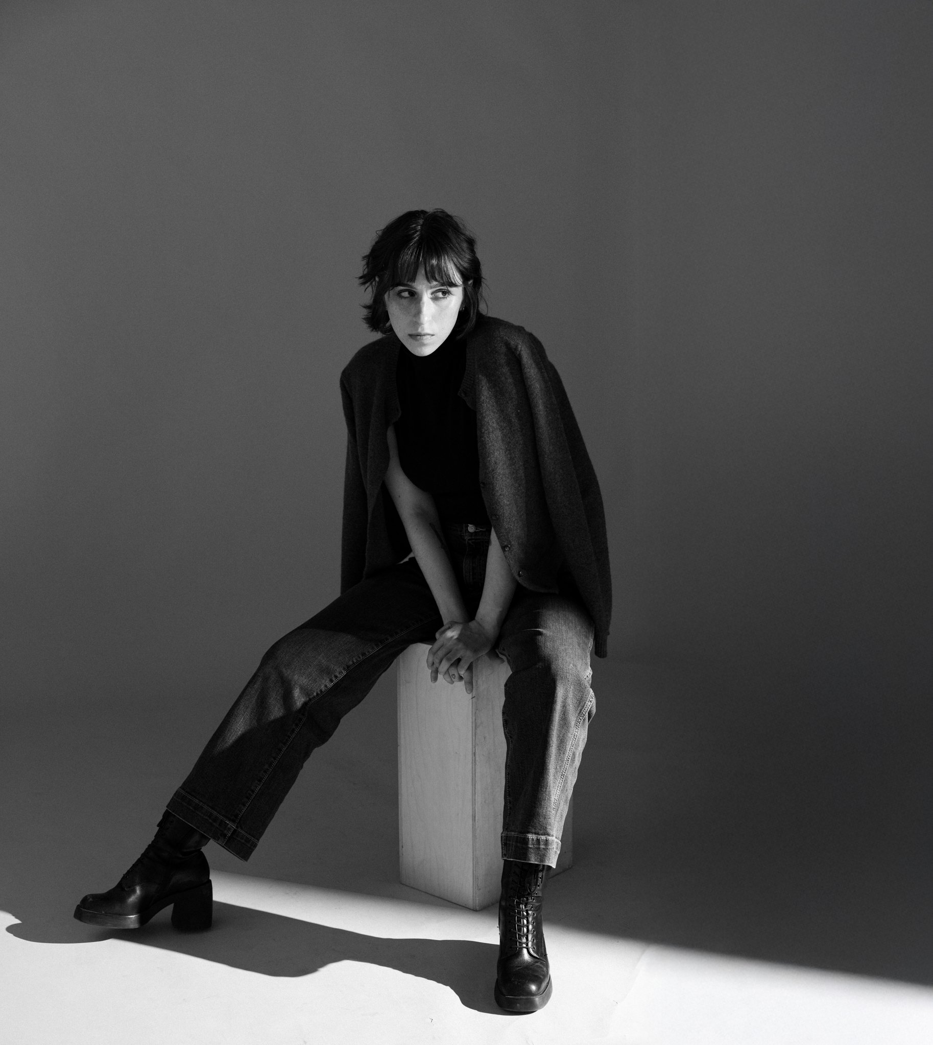 Black and white photo of Cleo Shine sitting on a high wooden stool, wearing a dark turtleneck, a textured coat, baggy jeans, and lace-up boots, with a plain gradient background.