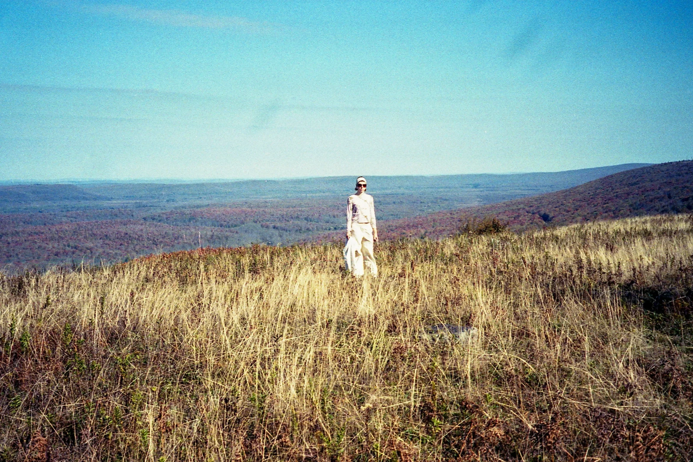 Woman standing in an open grassy field with distant hills and a clear blue sky.