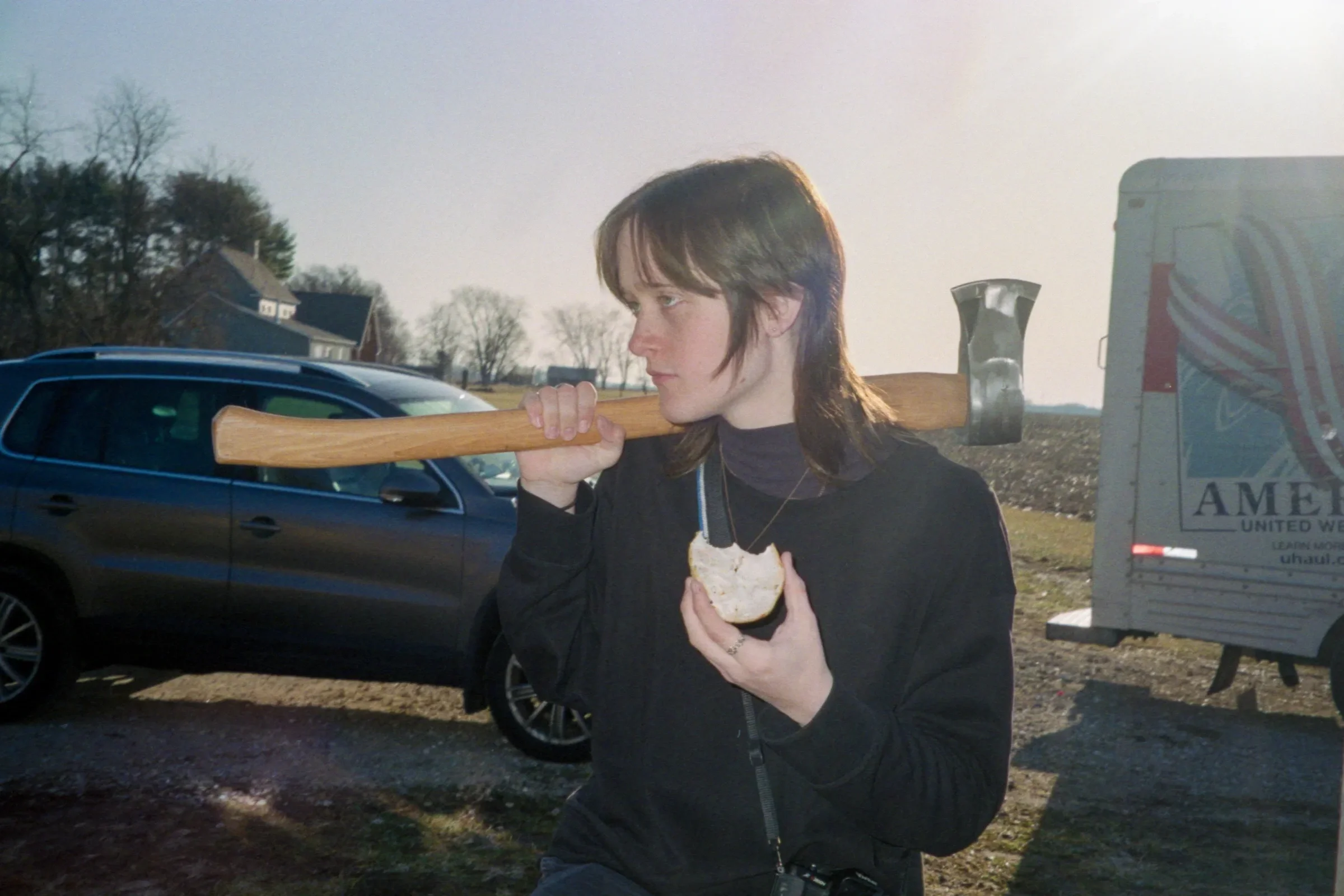 A person with shoulder-length brown hair, wearing a black jacket, is holding a sandwich in one hand and an axe over their shoulder with the other hand, standing outdoors near parked cars and a truck with a logo on it.