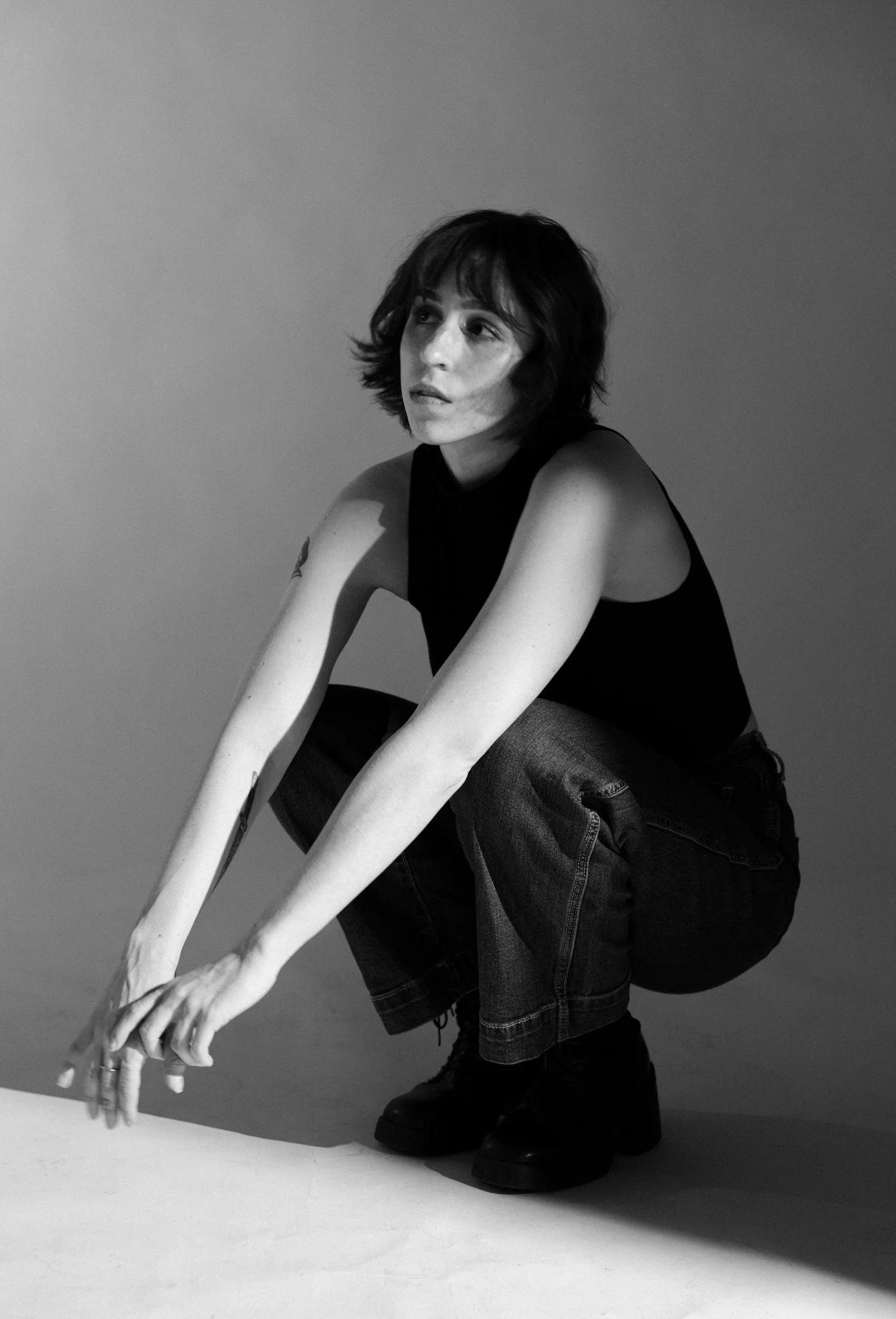 Cleo Shine with short, dark hair and light skin, wearing a sleeveless black top and jeans, is crouched down with their hands on the ground, looking upward to the left in a black-and-white photo.