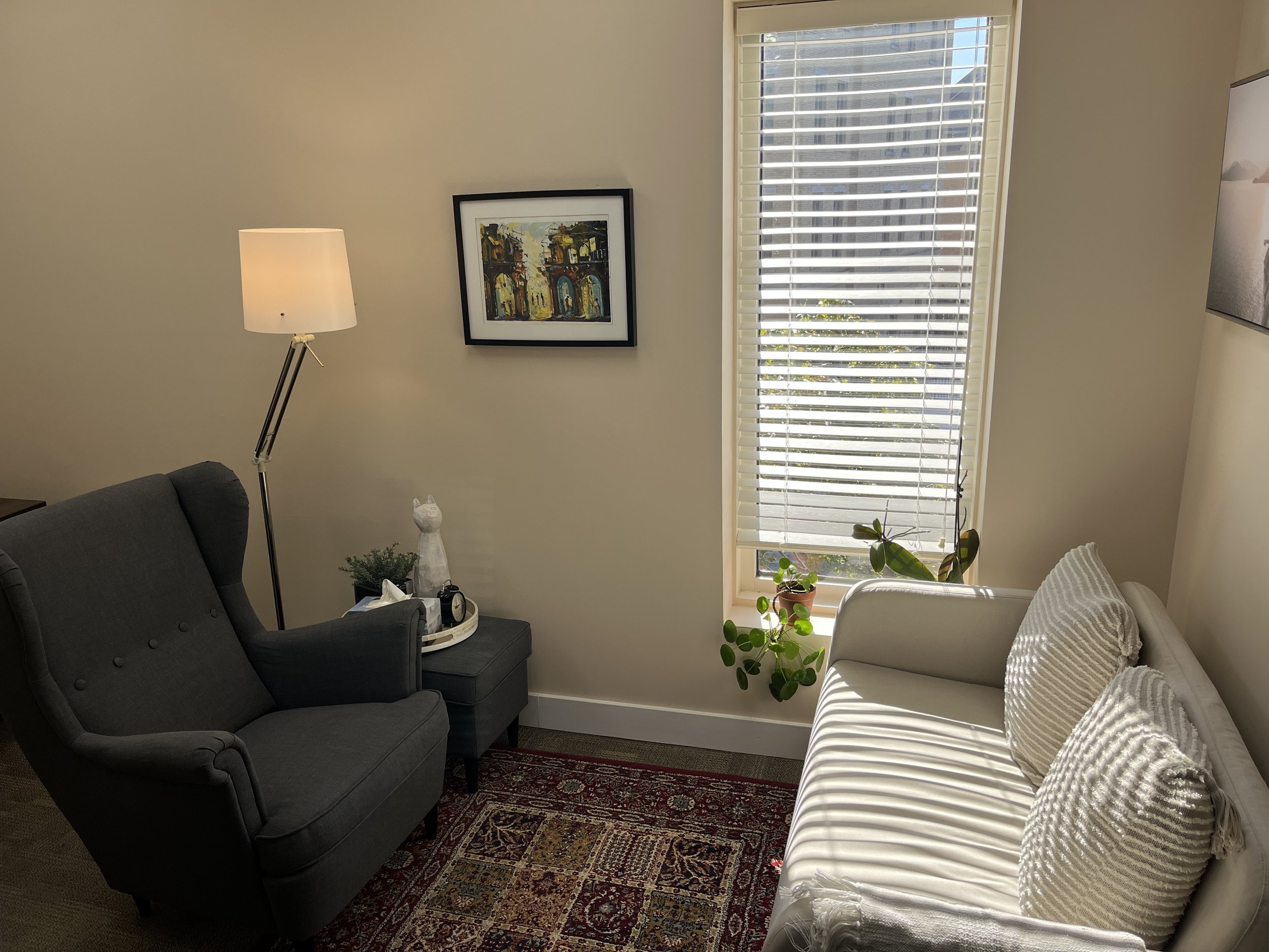 Cozy living room corner with gray armchair, white sofa with striped pillows, side table with owl decoration, table lamp, framed artwork, and a window with blinds letting in natural light.