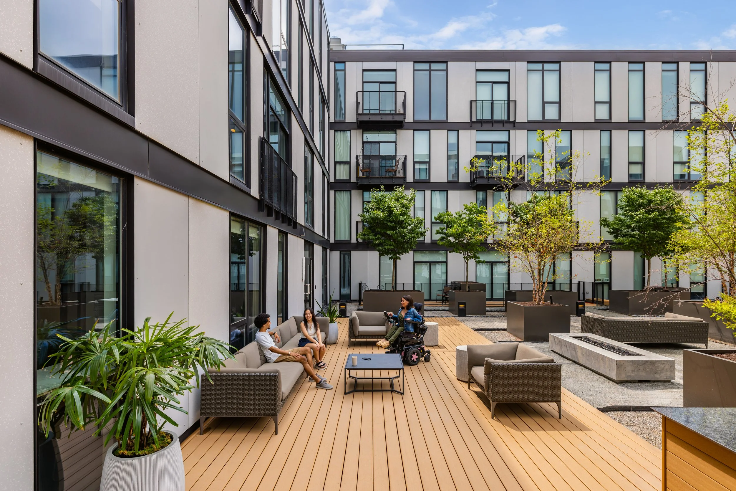 An exterior photo of a deck area at City Modern, showcasing a group talking; a man and woman sit on a couch across from a woman in a wheelchair.
