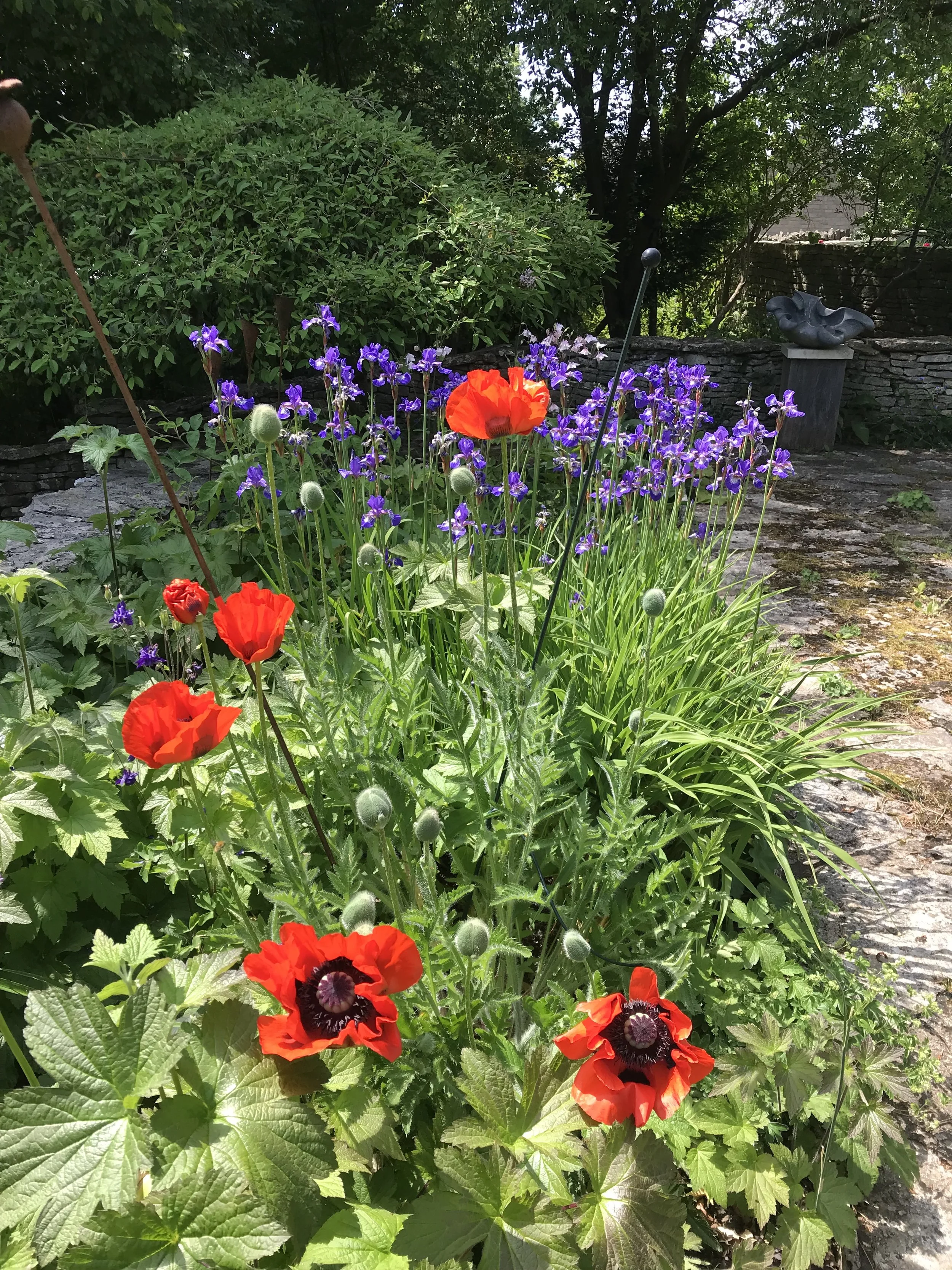 Garden with purple and red flowers, green plants, a stone path, a stone sculpture, and trees in the background.