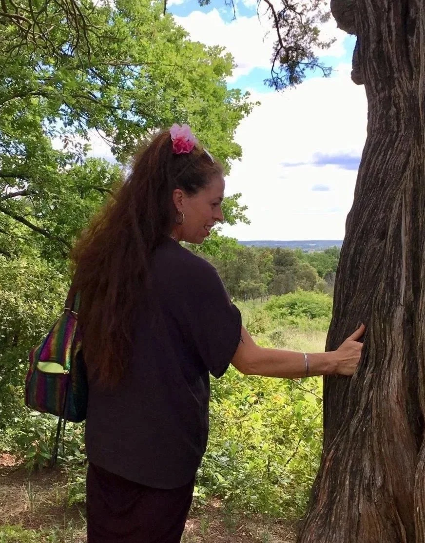 A woman with long brown hair, wearing a black shirt and a colorful backpack, is standing outside in a wooded area. She is smiling and touching the trunk of a large, old tree with textured bark. The background features green trees, a cloudy sky, and a distant landscape.