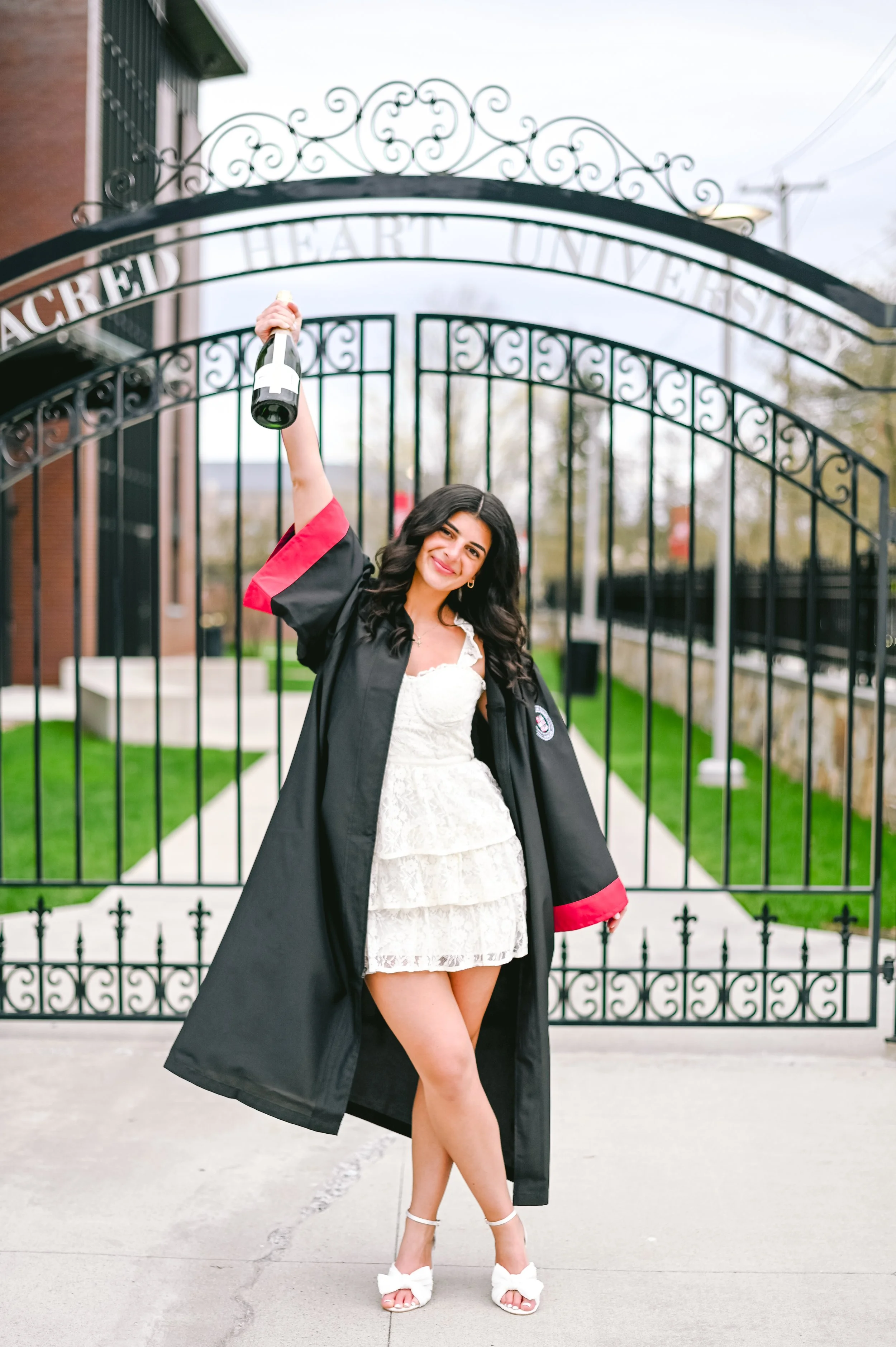 Senior graduate in a graduation gown standing in front of a gate, holding a diploma, smiling. She is having their senior grad photos taken at Sacred Heart University