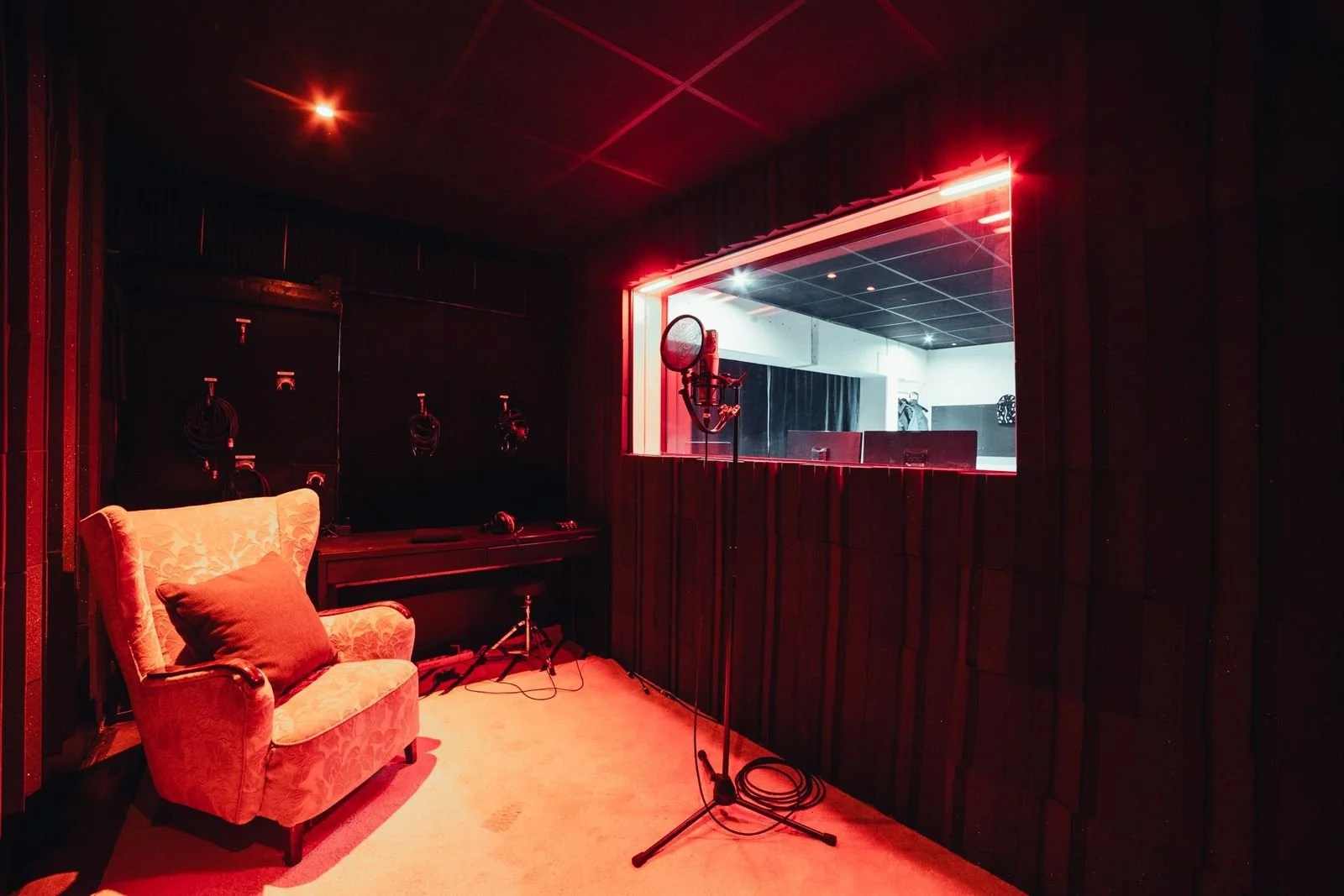 Sound recording studio with a vintage armchair, microphone, audio equipment, and a window looking into the control room, illuminated by red lighting.