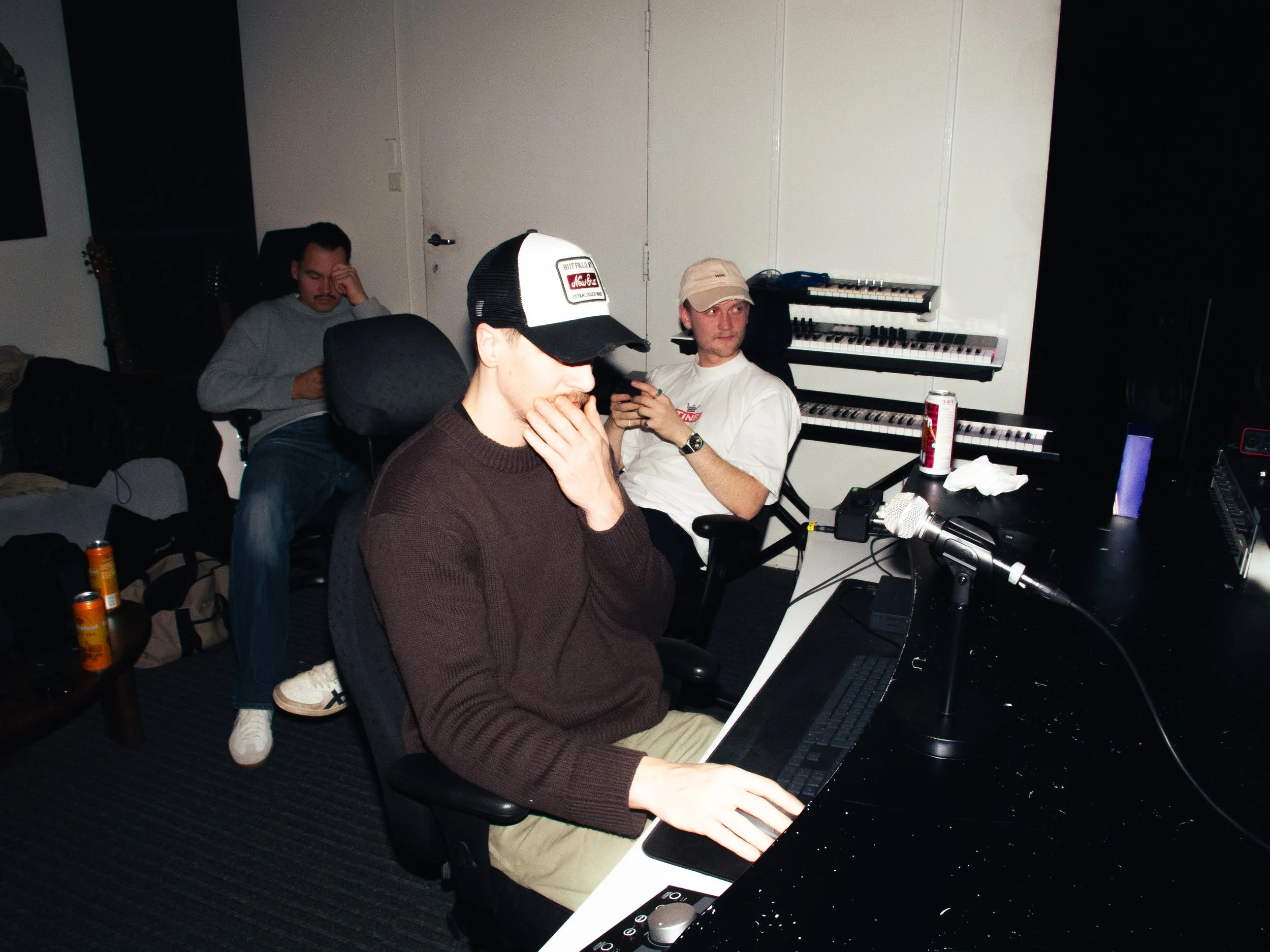 Three men in a music studio, one at a keyboard and two sitting, one with a laptop, with musical keyboards behind them.